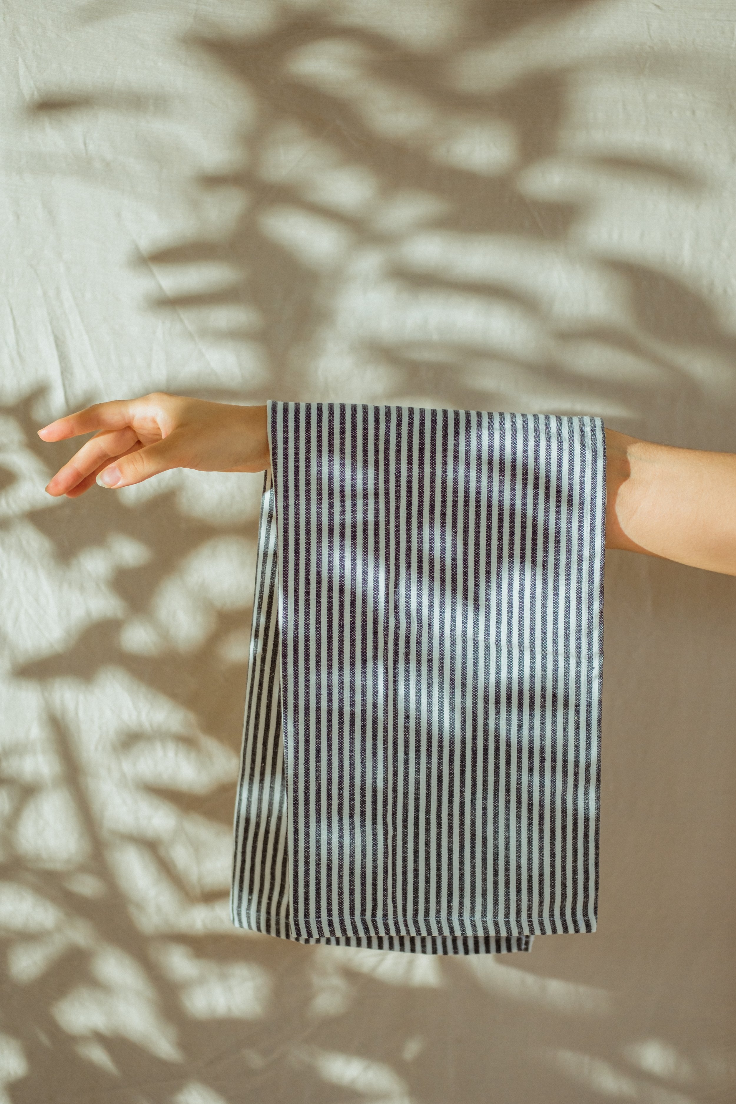 Close-up of a person's arm and hand resting on a fabric surface, with shadows cast by nearby leaves creating leaf-shaped patterns on the surface. The person is wearing a striped long-sleeve shirt.