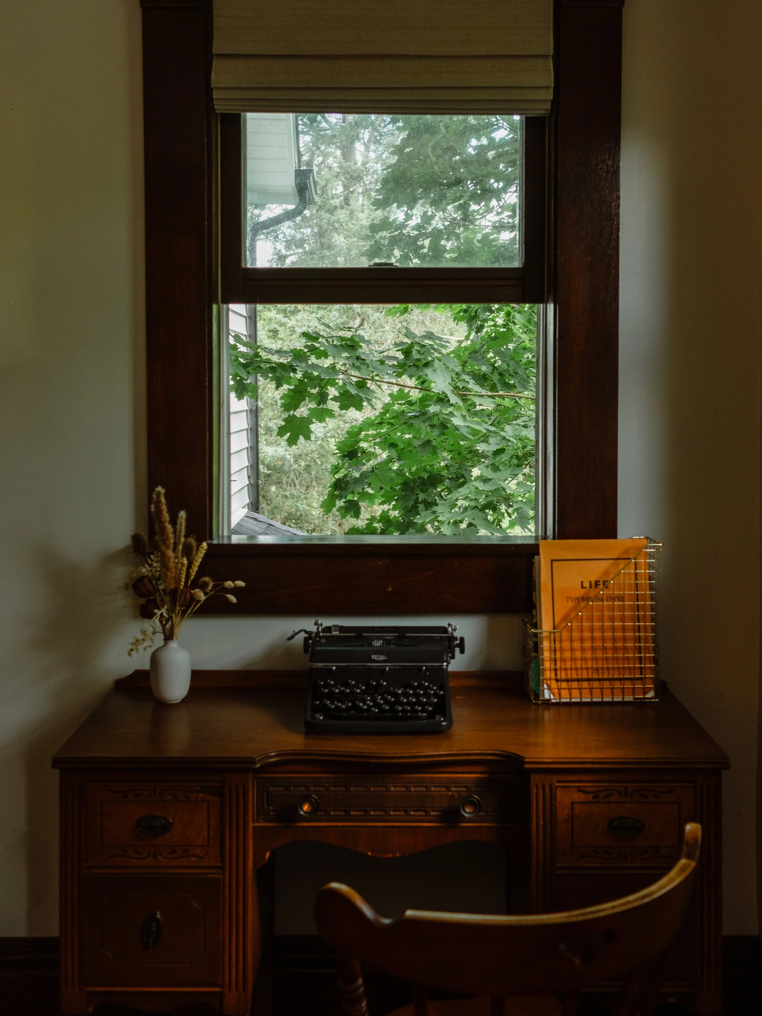 A window with a view of green leaves and trees outside, with a wooden desk below holding an antique typewriter, a white vase with dried flowers, and a yellow folder in a wire basket.