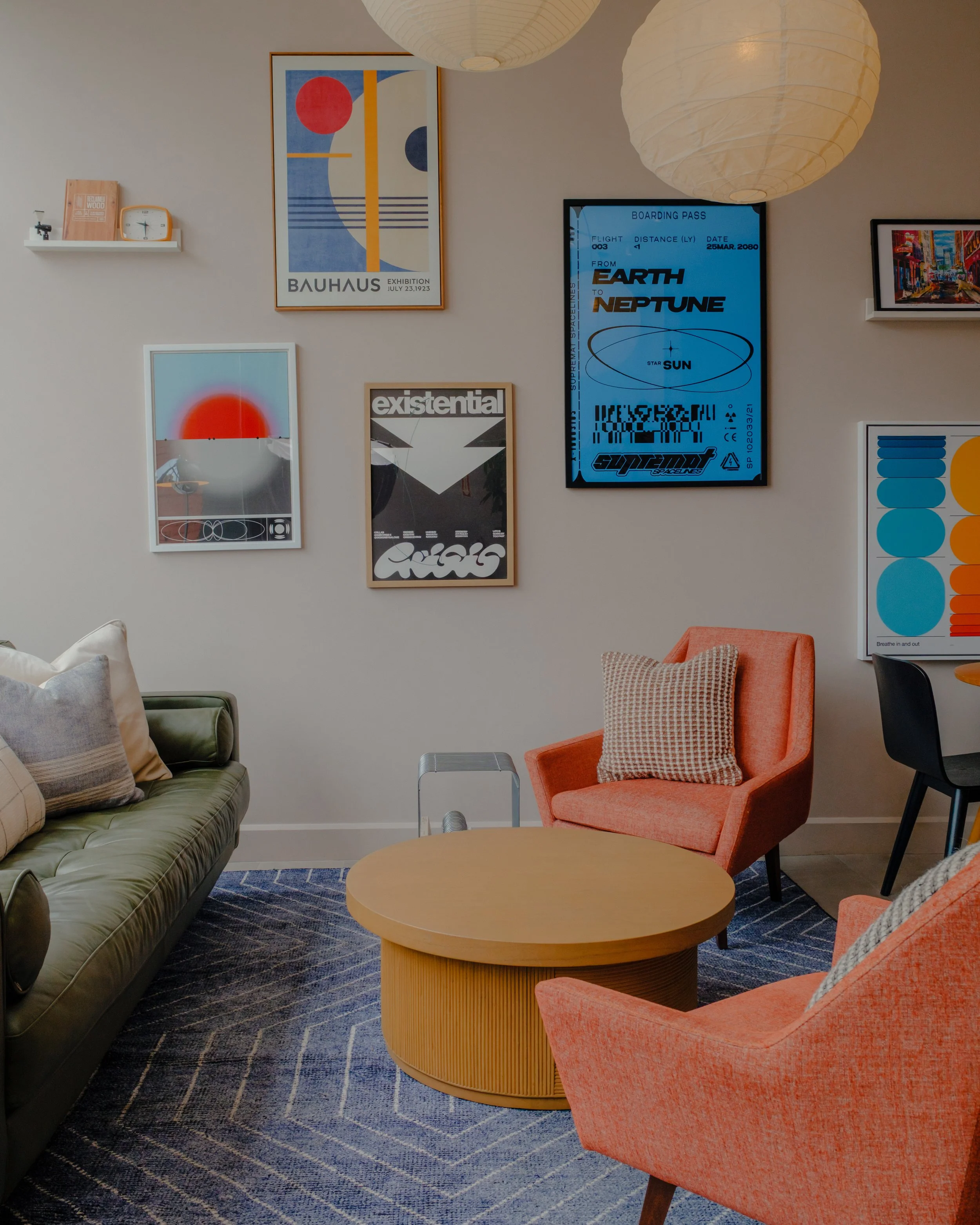 A cozy living room with a green sofa on the left, a peach armchair with a plaid pillow on the right, a round wooden coffee table in the center, and a blue patterned rug on the floor. The wall is decorated with framed artwork and a digital display. Tw