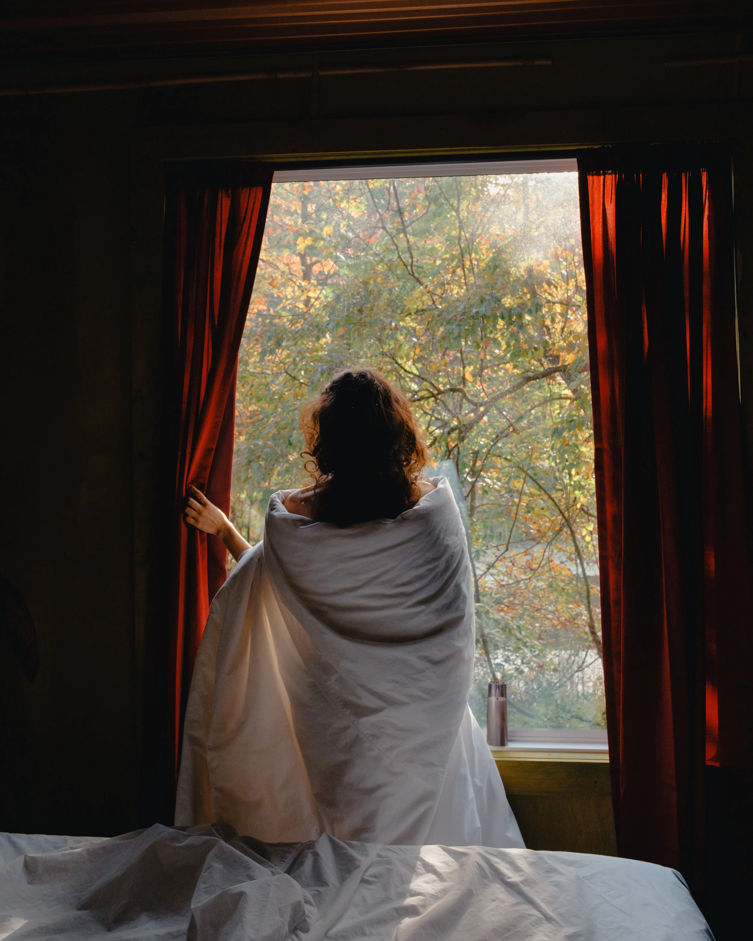 A person with curly hair, wearing a white robe, is sitting on a bed near a window and looking outside at autumn-colored trees. The window has red curtains pulled back, revealing a scenic view of fall foliage.