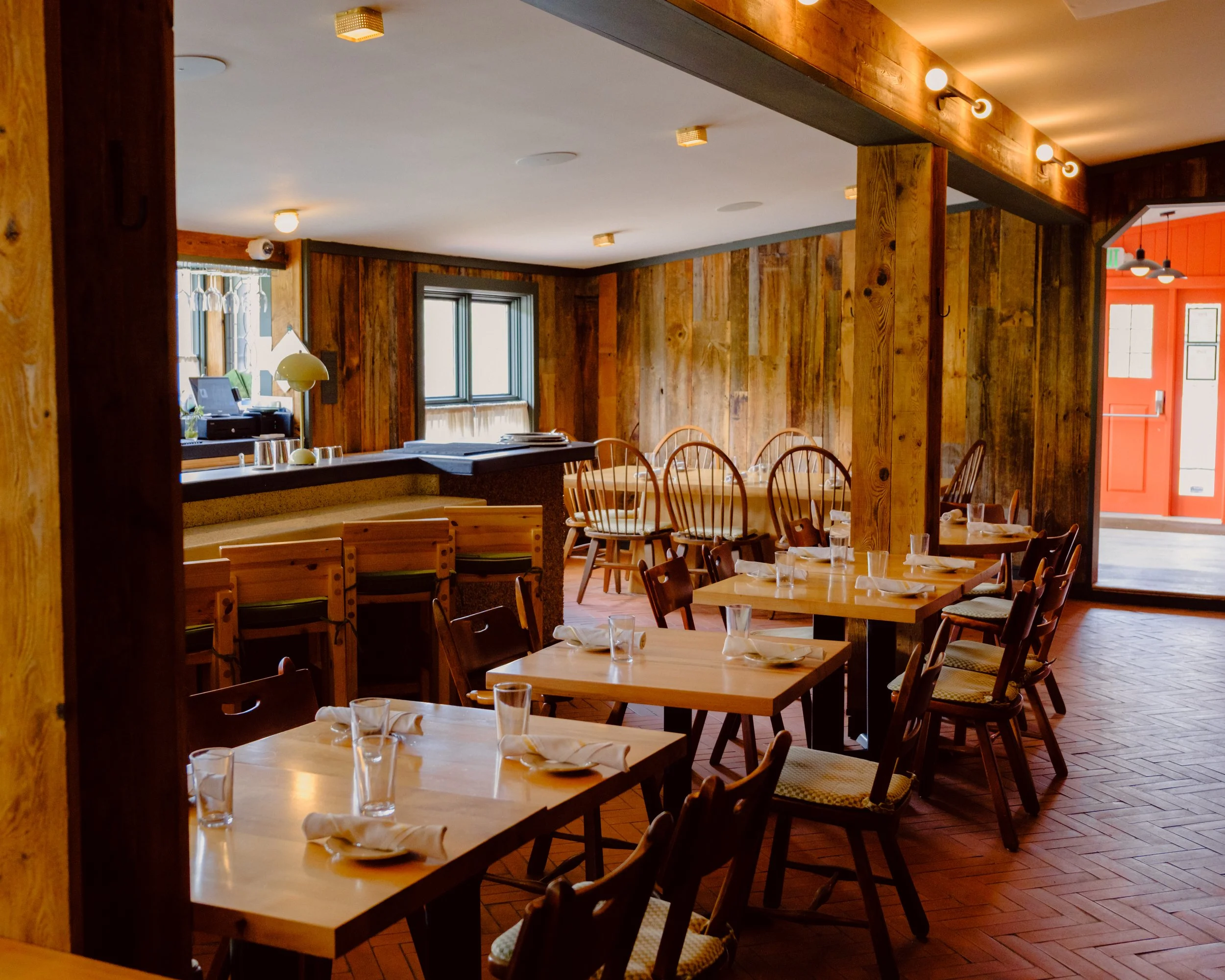 Interior of a cozy restaurant with wooden walls and tables set with glasses and napkins, with natural light coming through windows and warm lighting fixtures on the ceiling.
