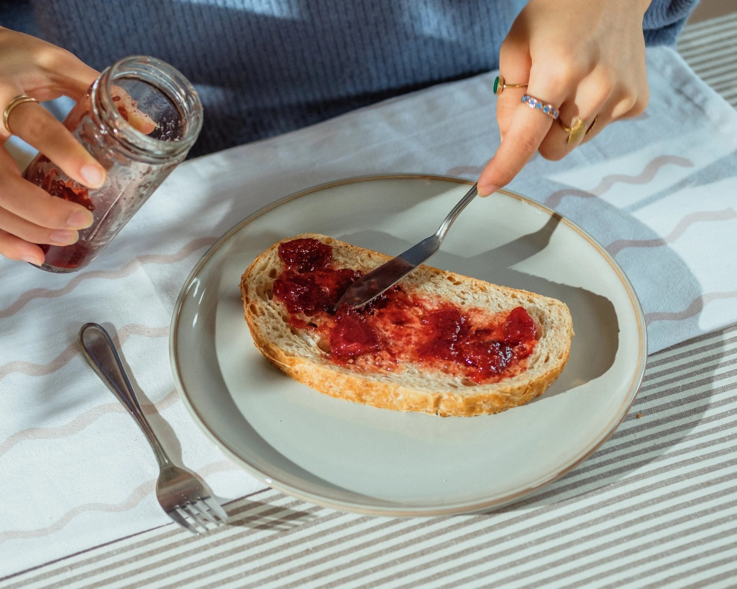 A slice of bread with red jam on a white plate, with a silver butter knife. A person is spreading jam on the bread using a spoon, and holding a jar of jam in their left hand. The person is wearing multiple rings, including a gold ring with a green st