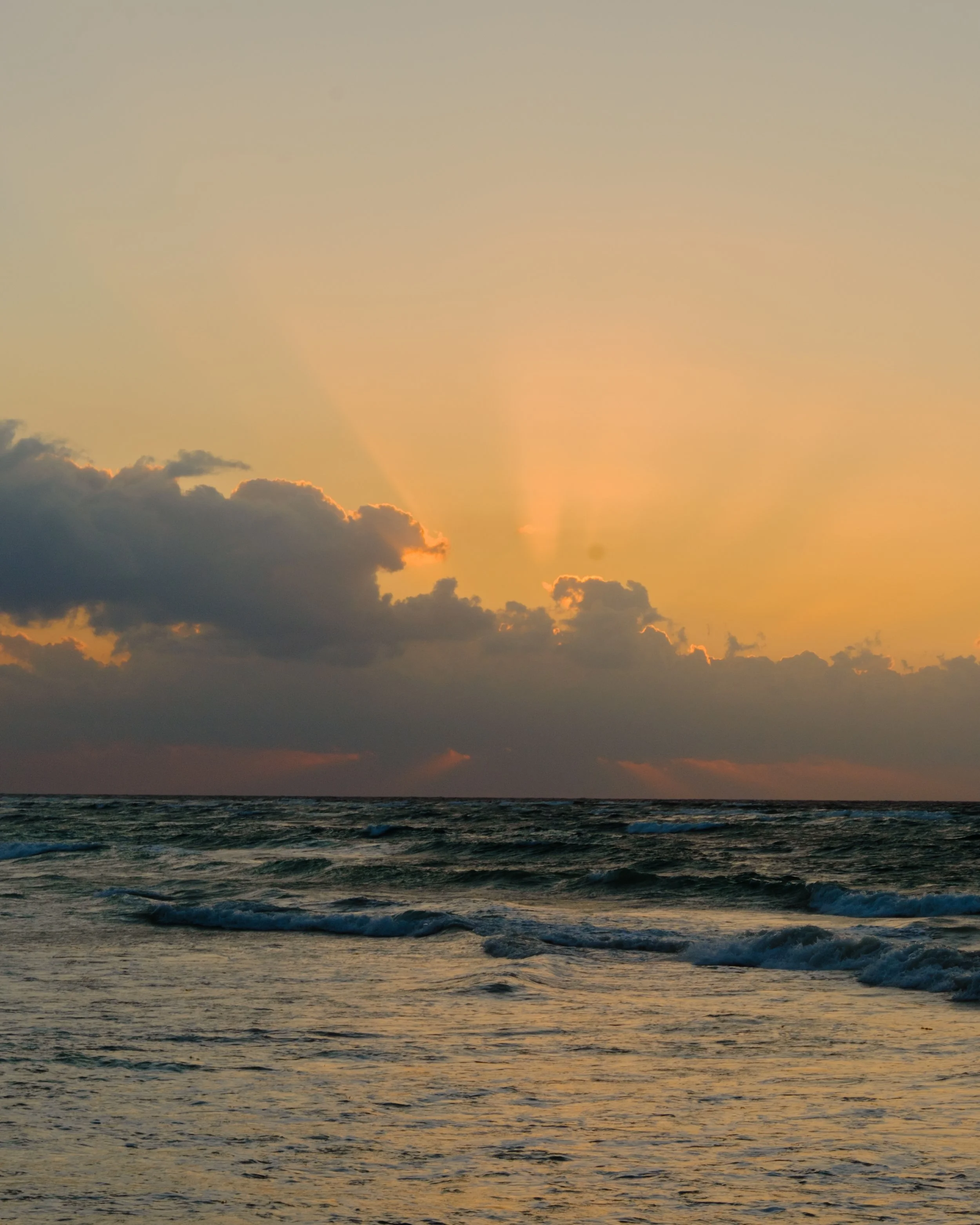 A sunset over the ocean with a sky filled with clouds and the sun setting beyond the horizon.