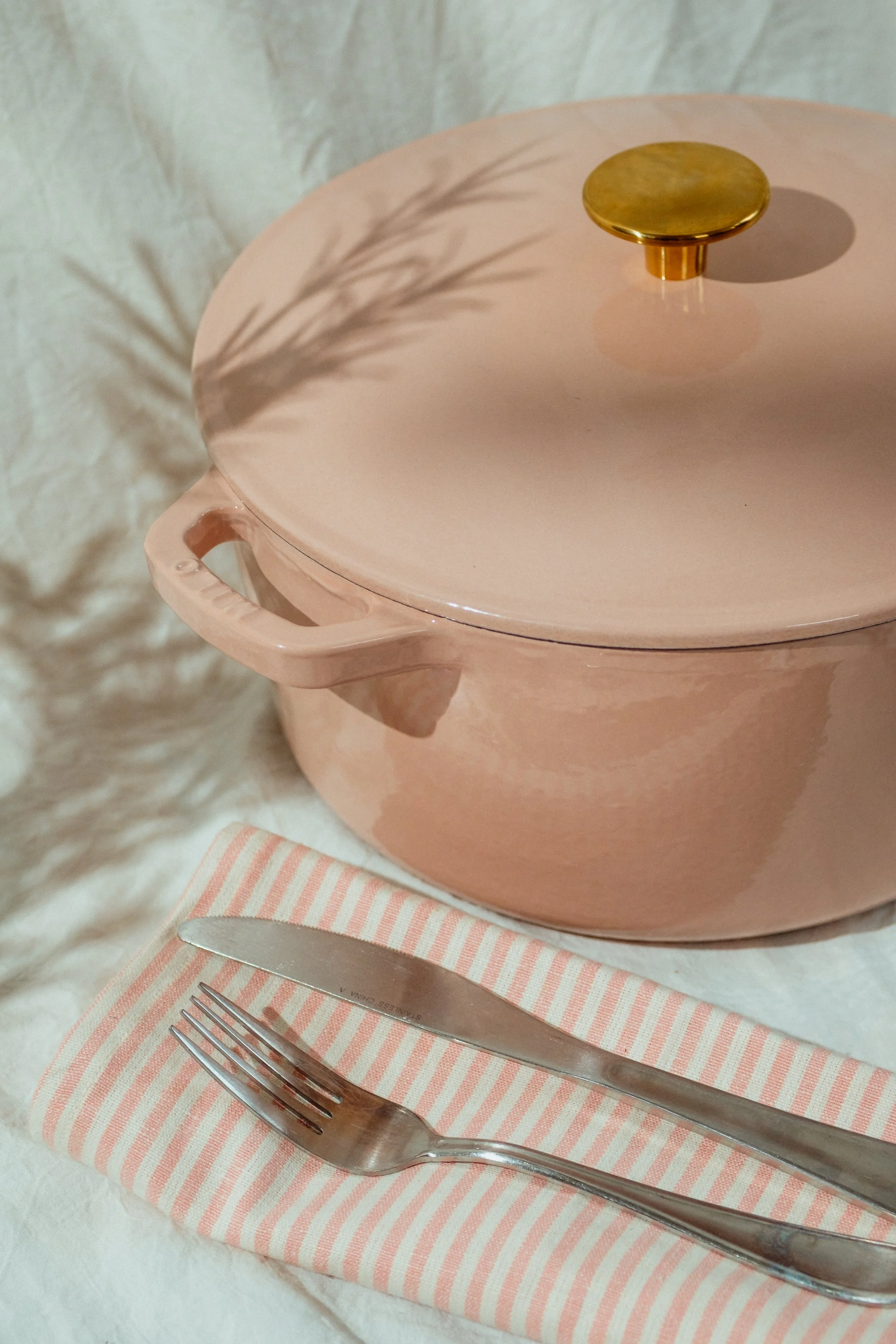 A pink ceramic pot with a lid decorated with a gold-colored handle, placed on a pink and white striped napkin. In front of the pot is a fork and knife on a white textured surface, with a shadow of a plant cast on the wall behind it's in a cozy settin