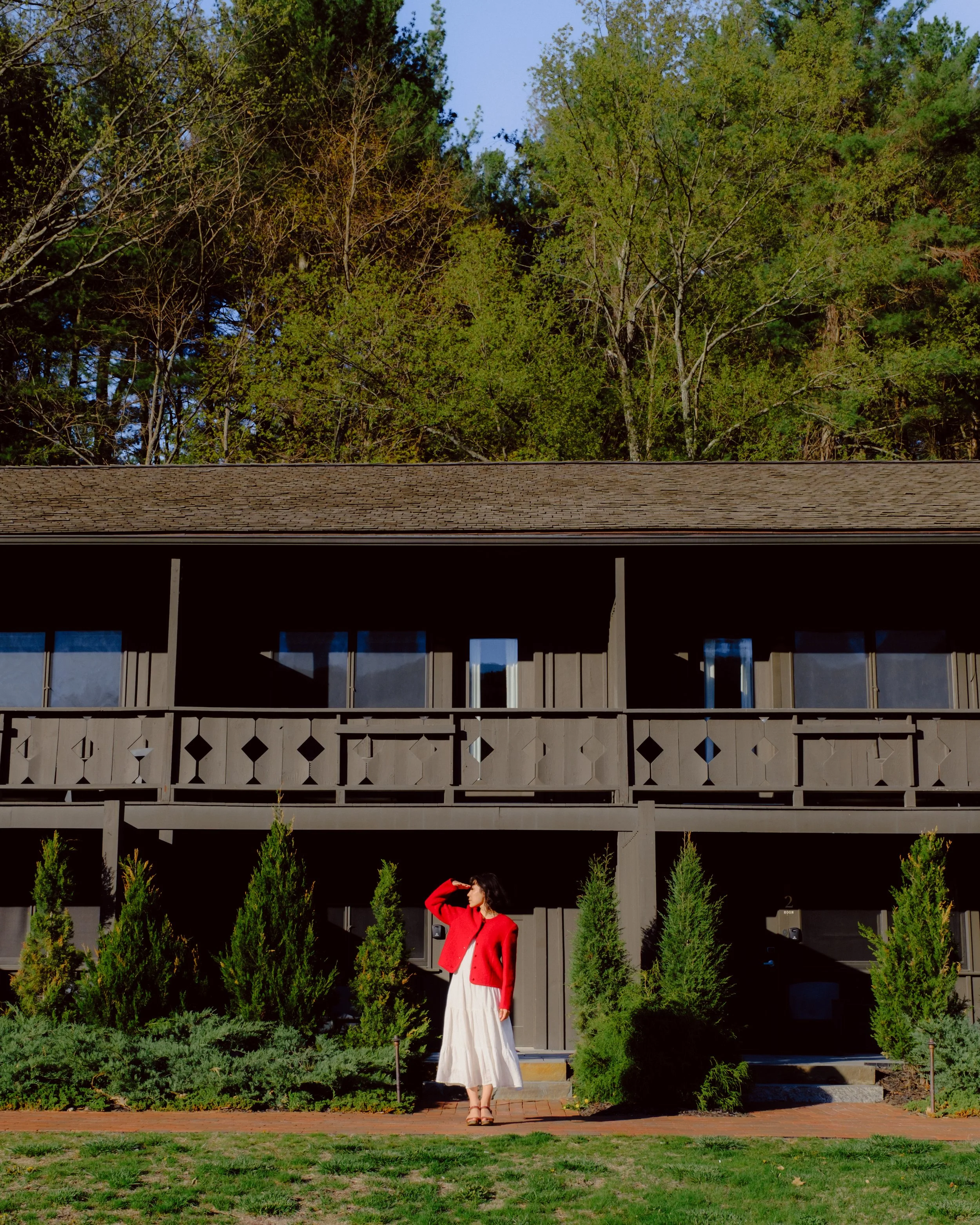 A woman with dark hair wearing a white dress and a red cardigan standing outdoors on a sunny day, framed by green bushes and trees, with a building and a clear blue sky in the background.