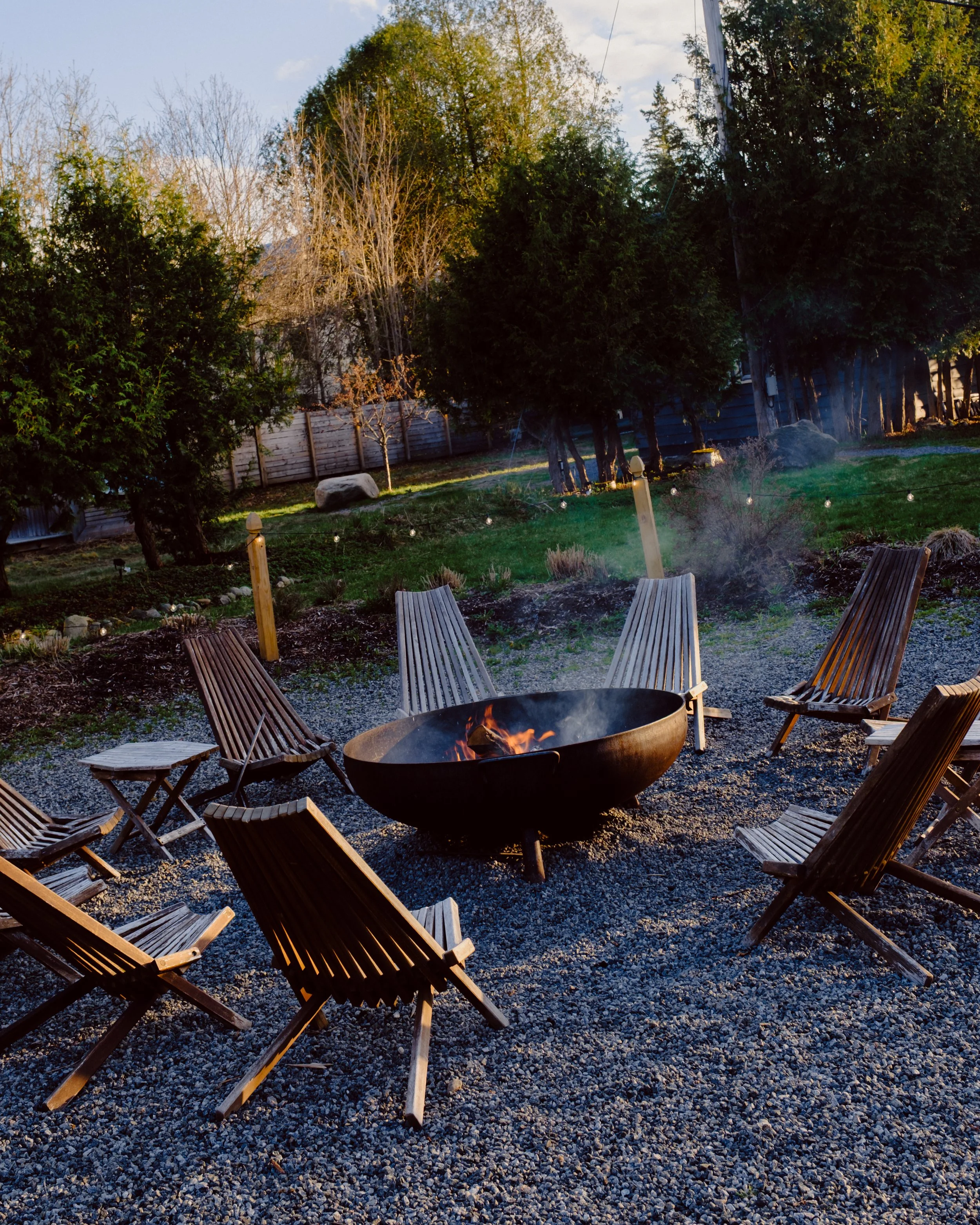 Backyard patio with wooden chairs arranged around a firepit, string lights, green grass, trees, and a wooden fence in the background, during late afternoon or early evening.