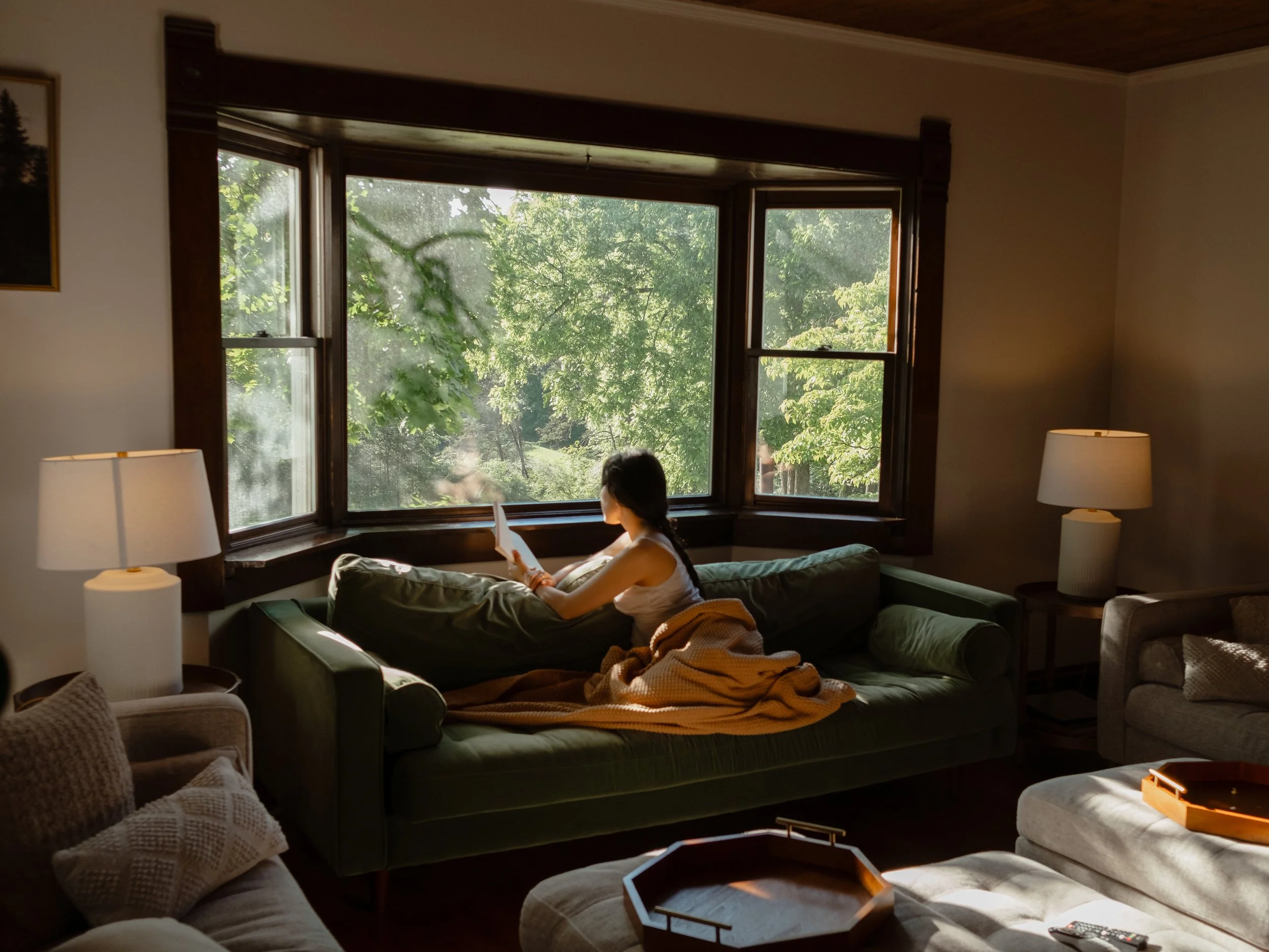 A woman sitting on a green couch reading a book by a large bay window showing green trees outside, with a beige lamp on a side table and pillows on the couch.