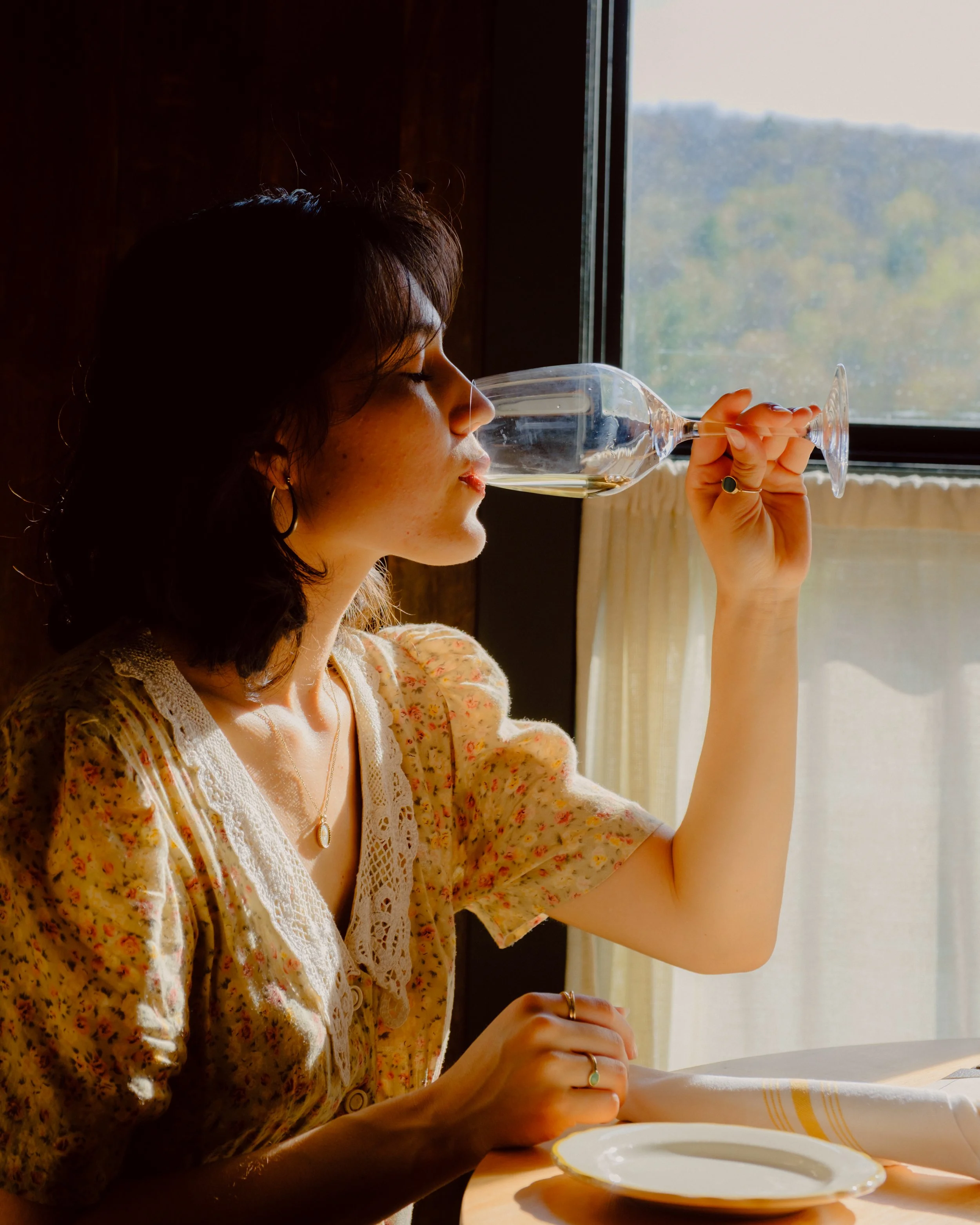 A woman with dark hair, wearing earrings and jewelry, sits at a table with a plate and a cloth. She is holding a wine glass close to her lips, with sunlight coming through a window behind her, casting a warm glow on her face and the table.