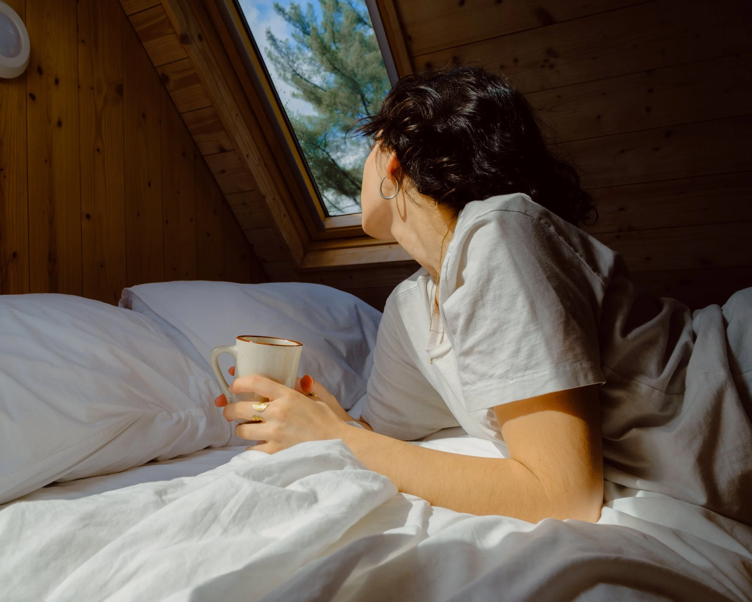 A person with curly dark hair and hoop earrings is sitting on a bed, holding a white mug, and looking out a window at the trees outside.