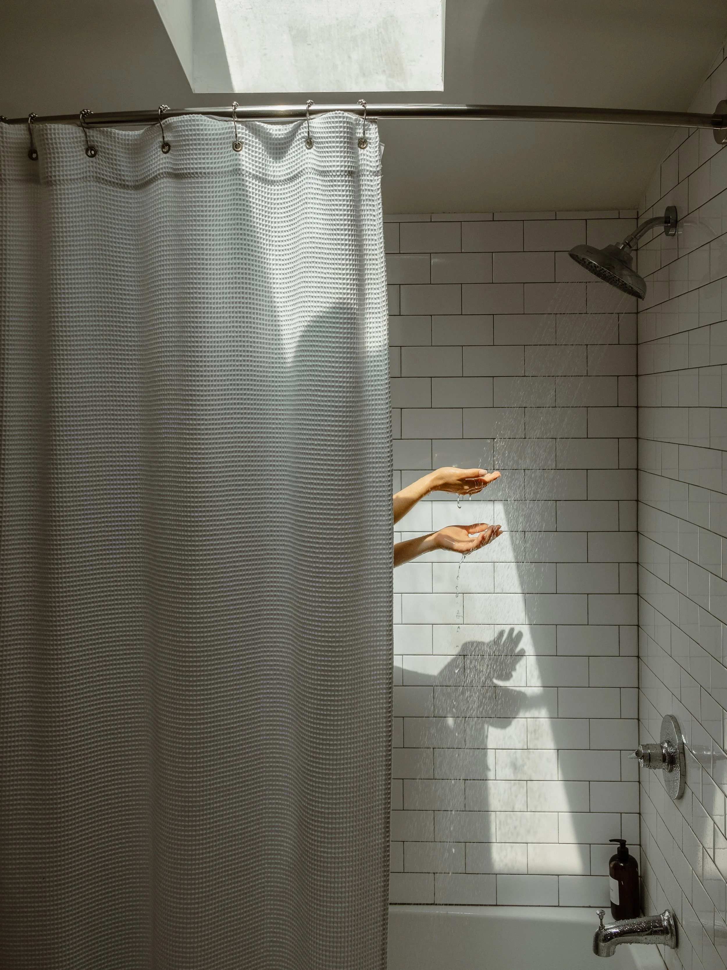 Inside a bathroom shower, with white tiled walls, a gray shower curtain on a rod, and a person's hands reaching out from behind the curtain with water running from the showerhead.