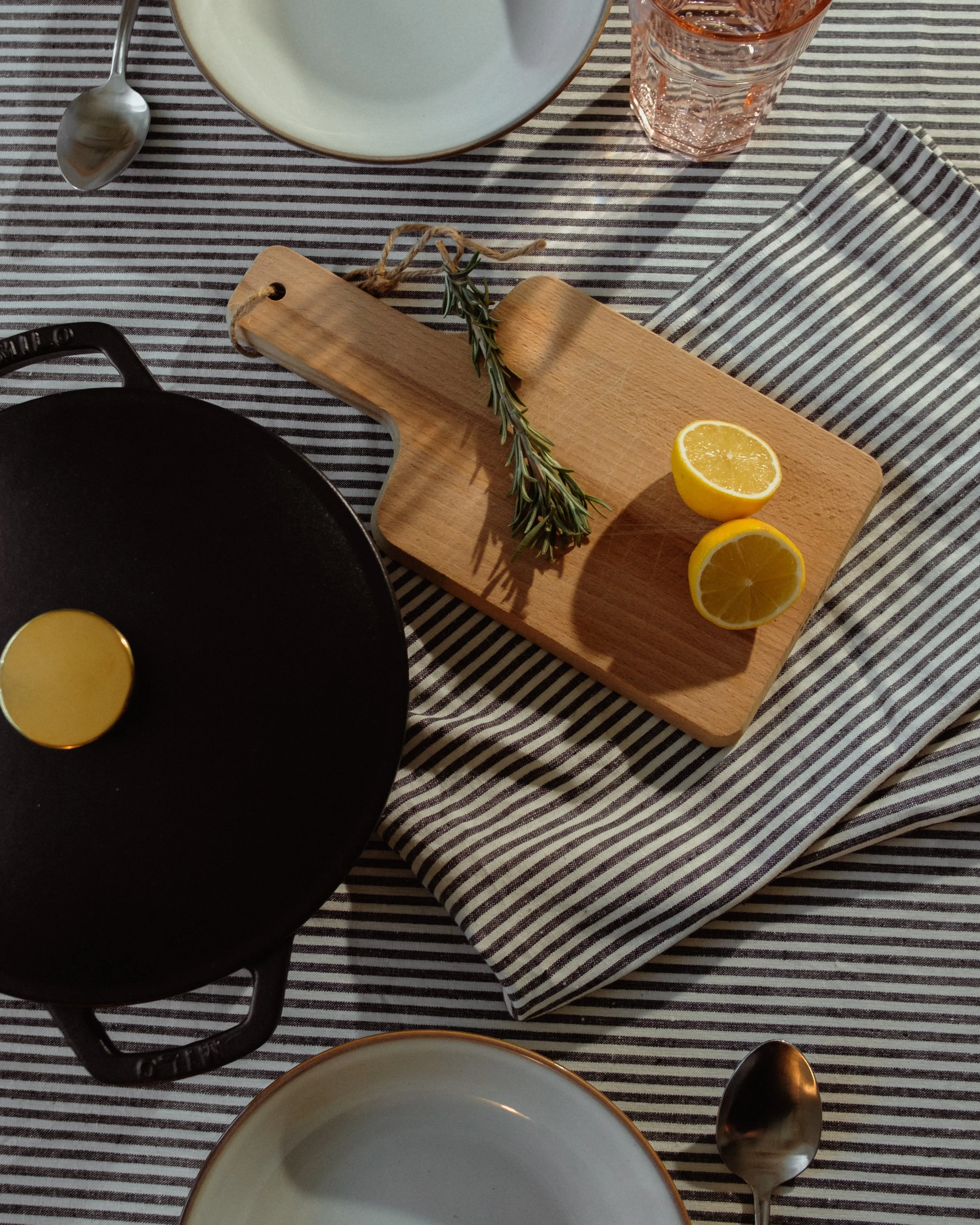 A dining table set with striped tablecloth, empty plates, silverware, a glass of water, lemon halves, a cutting board with rosemary and lemon, and a cast iron skillet.