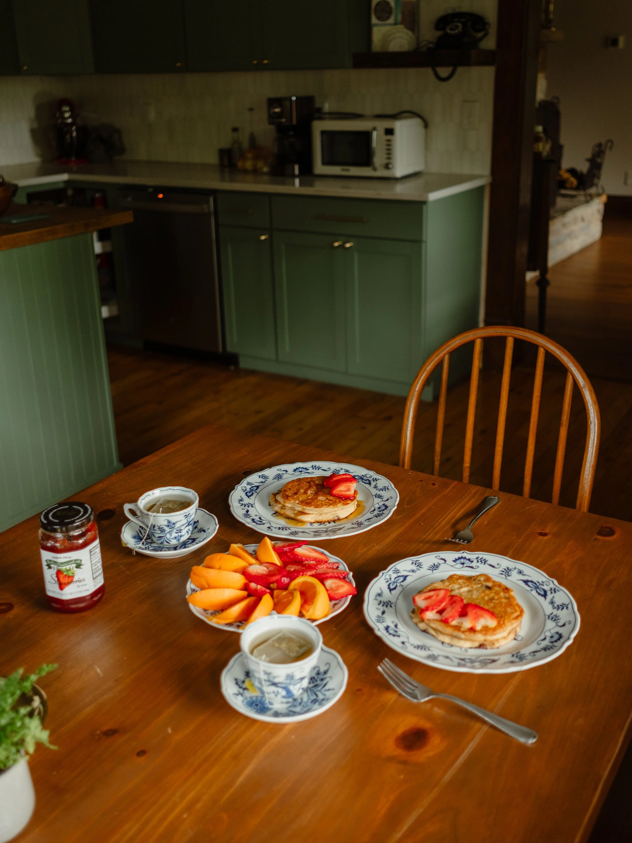 A breakfast table with pancakes topped with strawberries, a fruit salad, cups of coffee, jam, and utensils in a cozy kitchen setting.