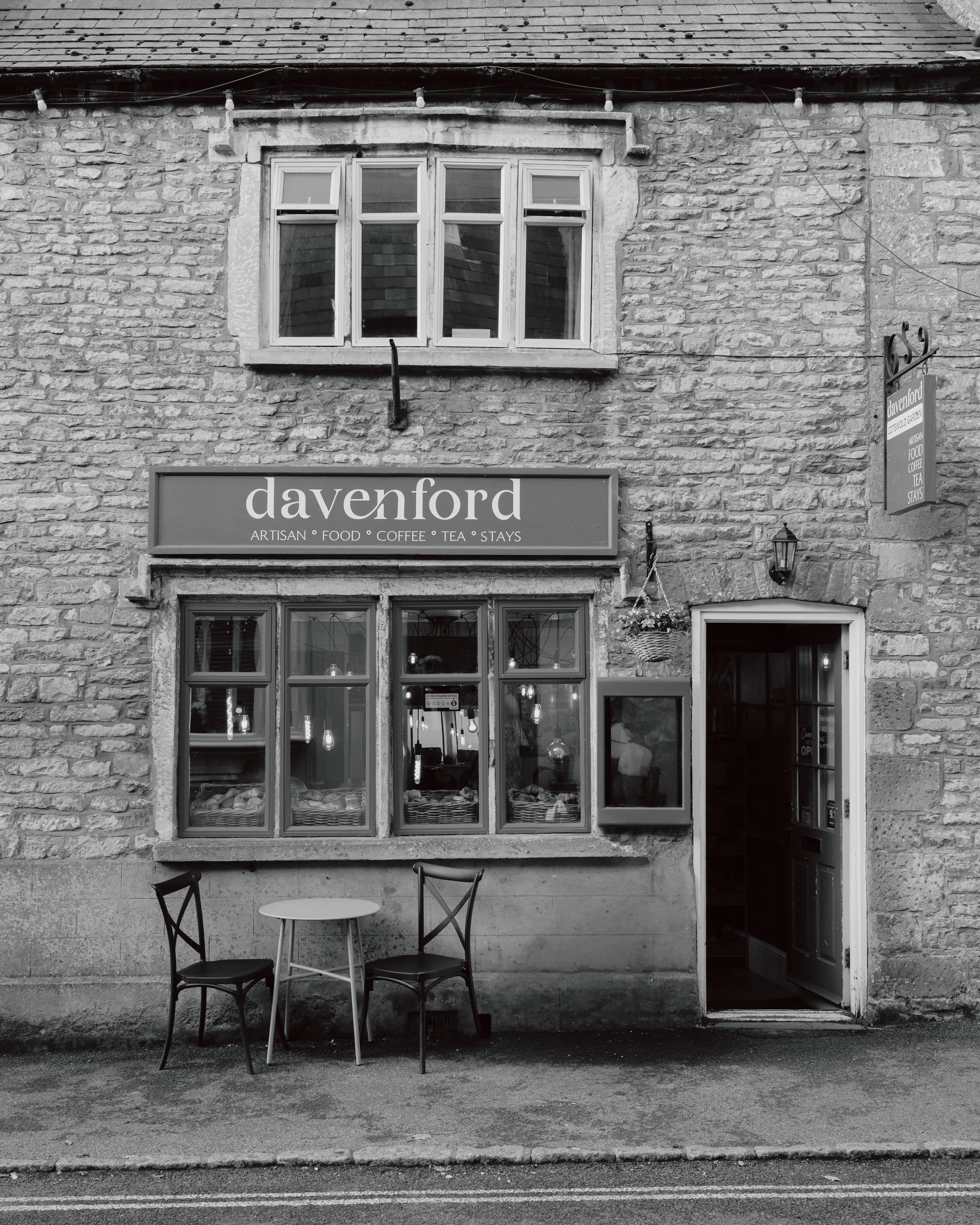 The exterior of a café or restaurant called 'davenford' with a sign reading 'artisan, food, coffee, tea, stays'. The storefront has a large window and an open door, with three chairs and a small round table in front of it. The building has a brick facade and a hanging plant on the side.