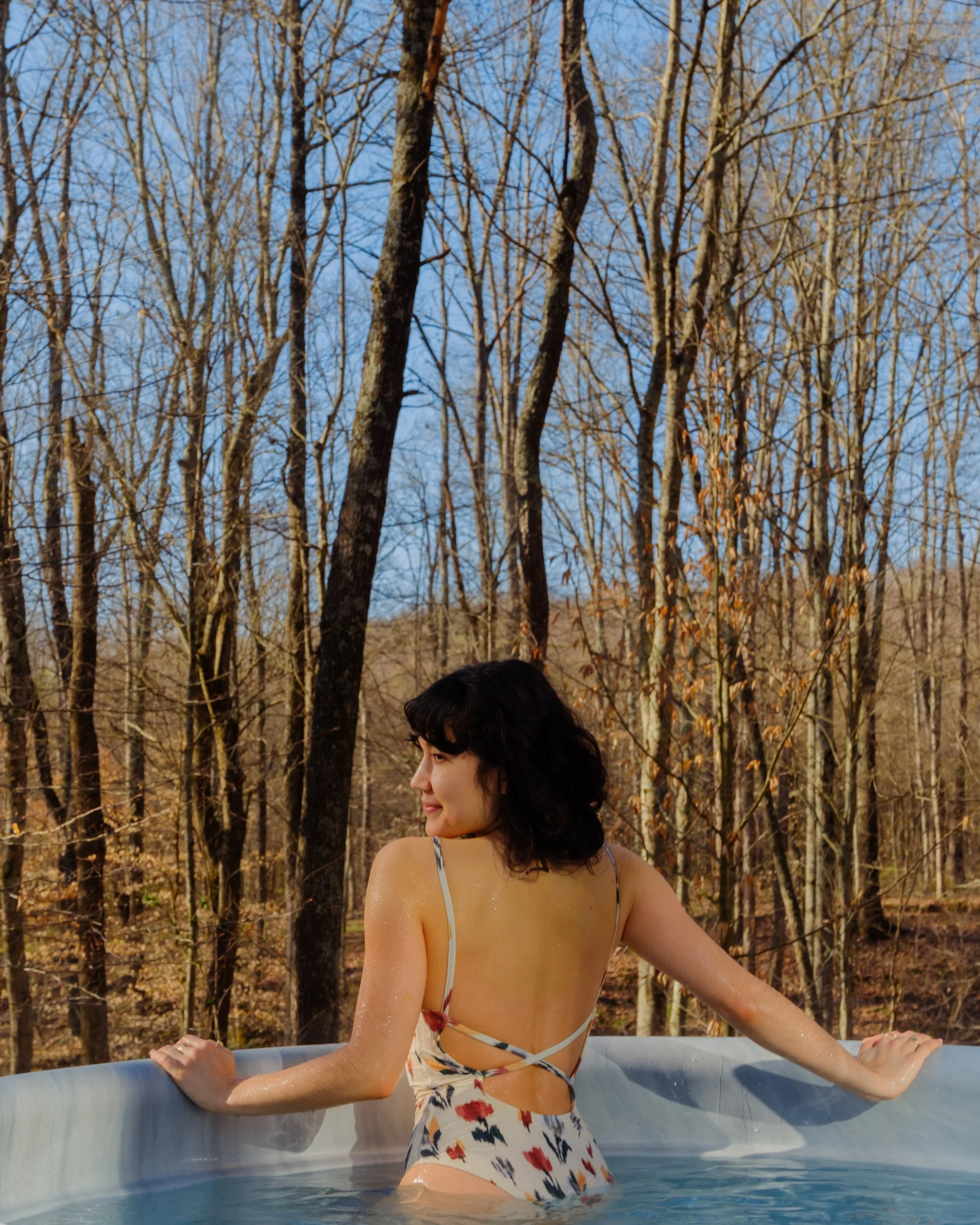 A woman is in a hot tub outdoors, wearing a floral swimsuit, with a backdrop of leafless trees and a blue sky.