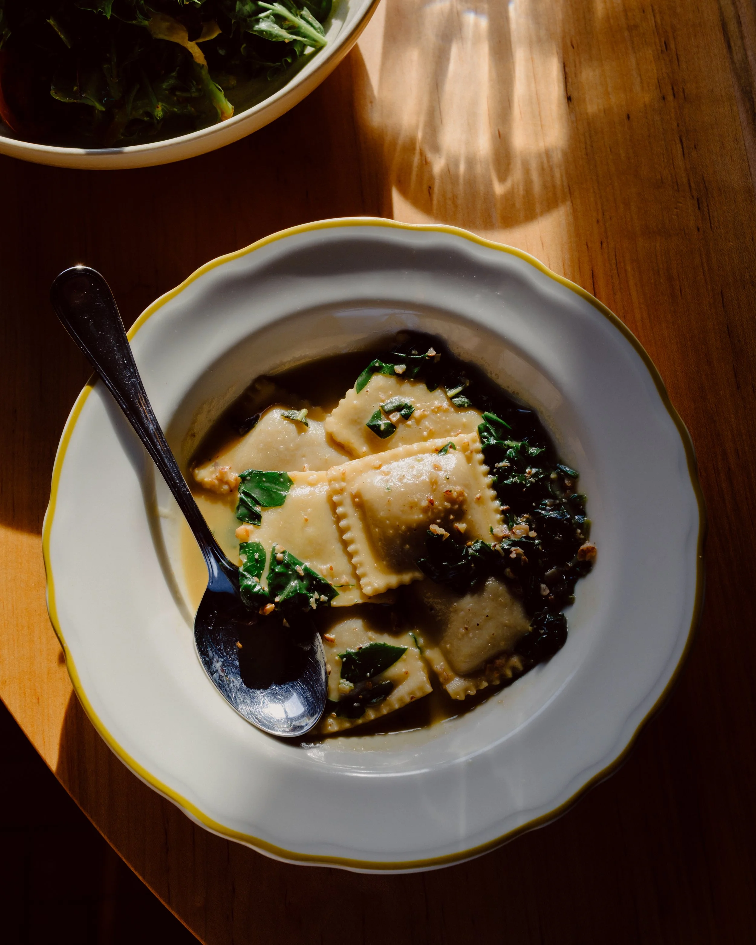 A bowl of beef noodle soup with sliced beef, green vegetables, and dark broth, served on a white plate with a black spoon, on a wooden table. In the background, there is a bowl of greens in a white bowl.
