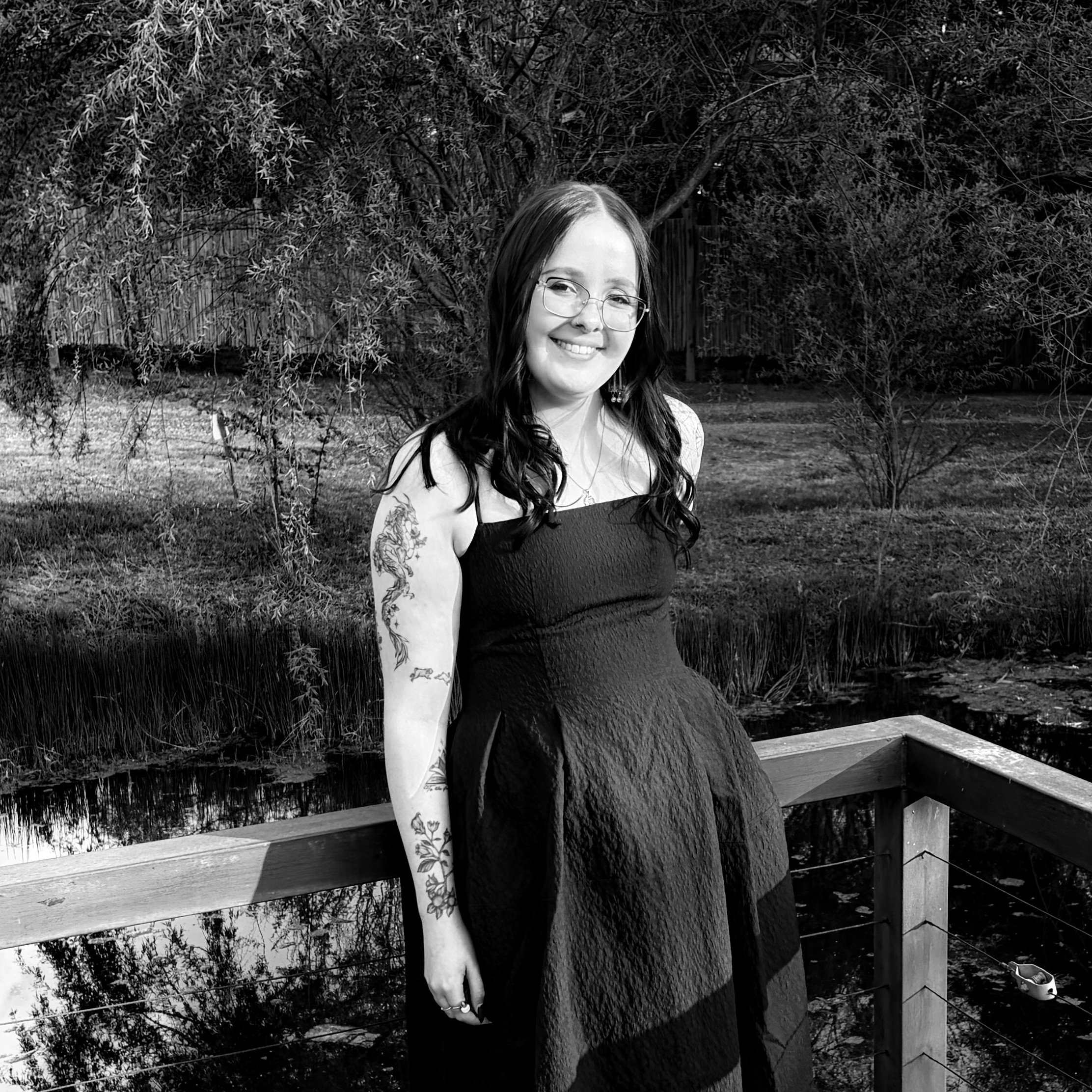 A smiling woman with glasses and long dark hair standing outdoors near a wooden fence, with trees and bushes in the background.