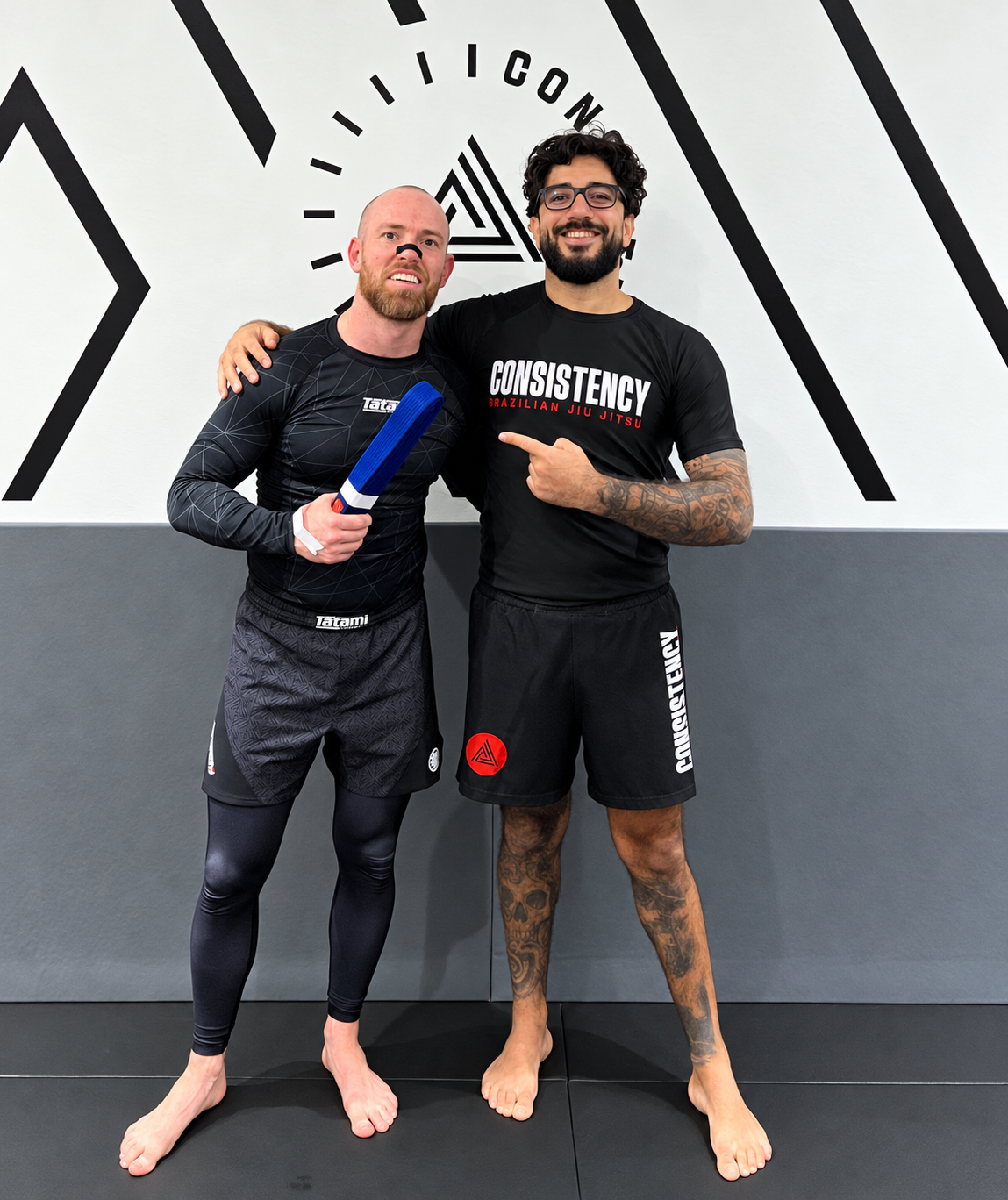 Two men in martial arts training gear standing together in a gym, smiling, with one holding a blue belt. The man on the left is wearing a black rash guard and shorts, and the man on the right has tattoos, a black T-shirt with white and red text, and black shorts. They are standing on a mat in front of a wall with black and white martial arts logos.