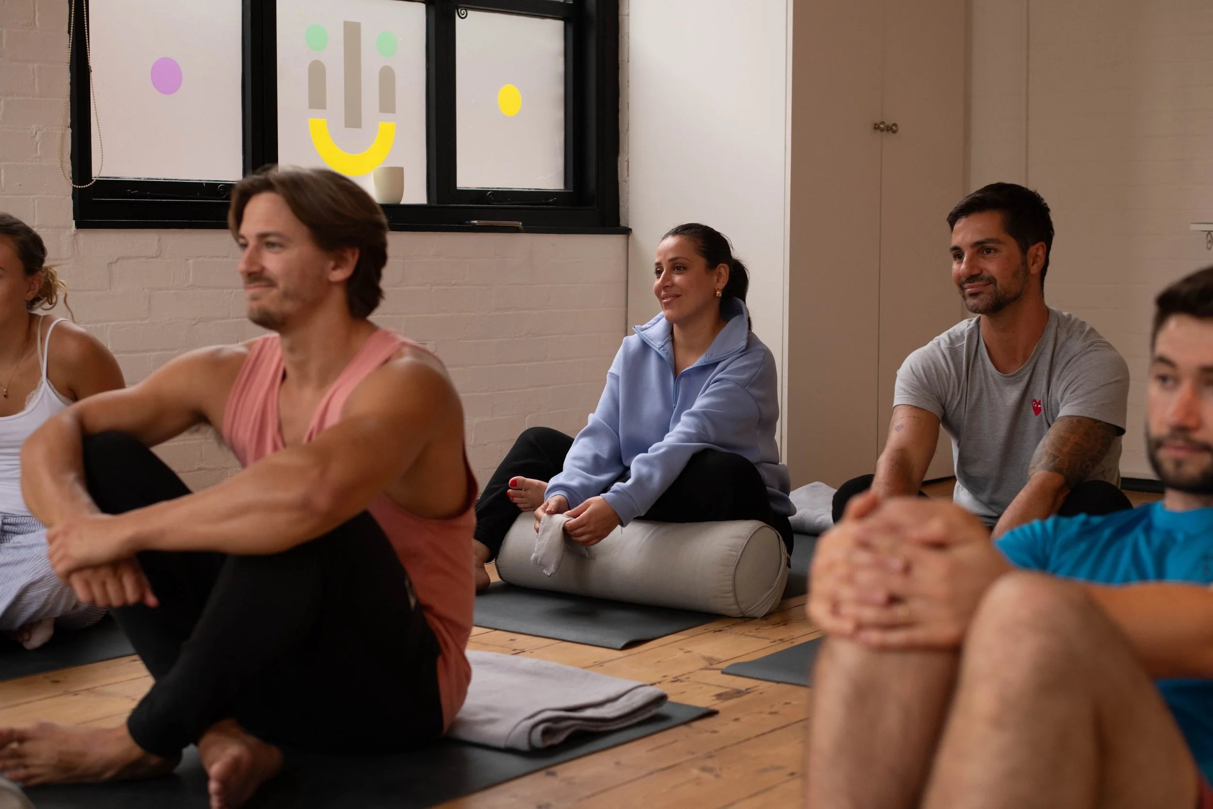 Group of people participating in a yoga or meditation class indoors, sitting on mats with some sitting on a cushion, with a window in the background decorated with colorful smiley face stickers.