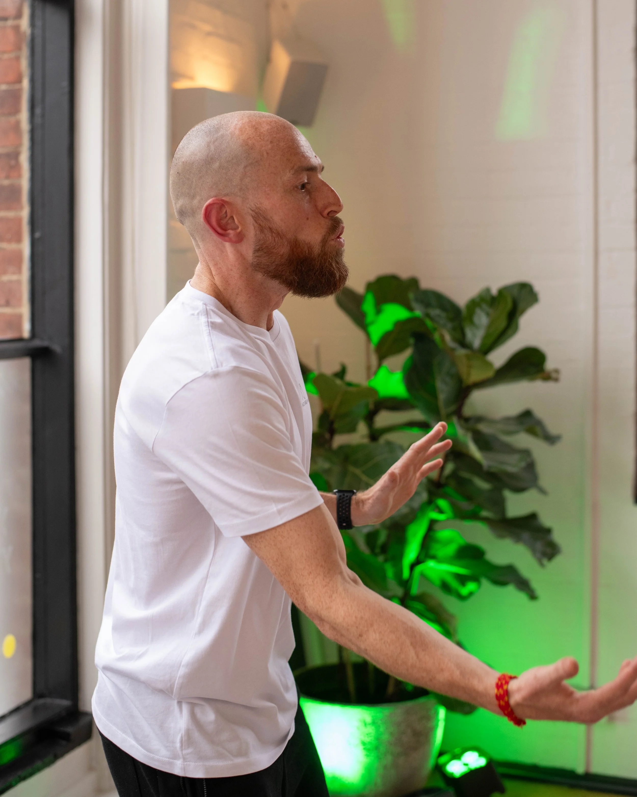 A man with a shaved head and beard wearing a white shirt and black pants, standing indoors with a green-lit plant in the background, appears to be meditating or practicing mindfulness with eyes closed and hands extended forward.