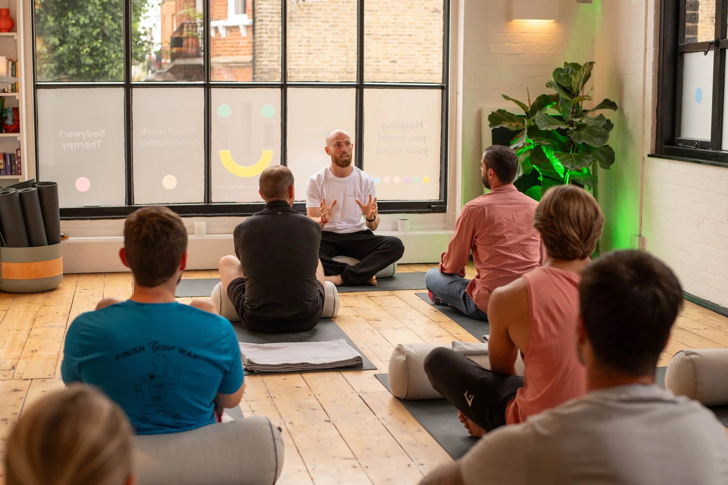 A man leading a meditation or yoga class with several participants sitting on mats and cushions in a bright studio with large windows, indoor plants, and bookshelves.