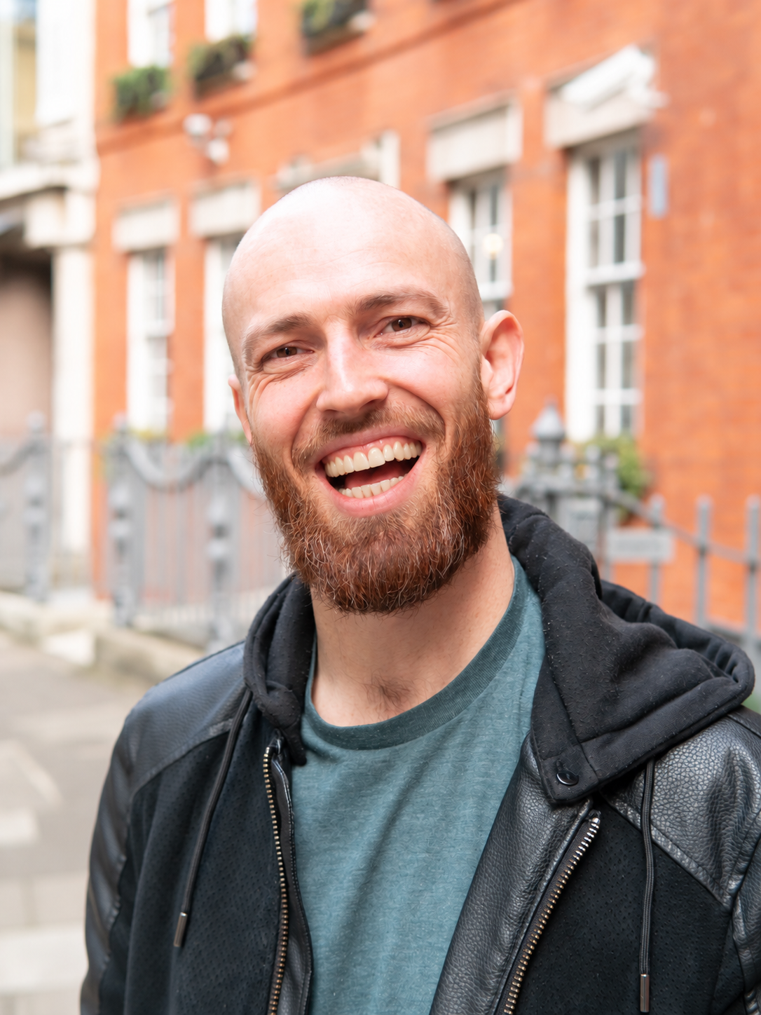A smiling man with a beard and bald head outdoors in front of a brick building.