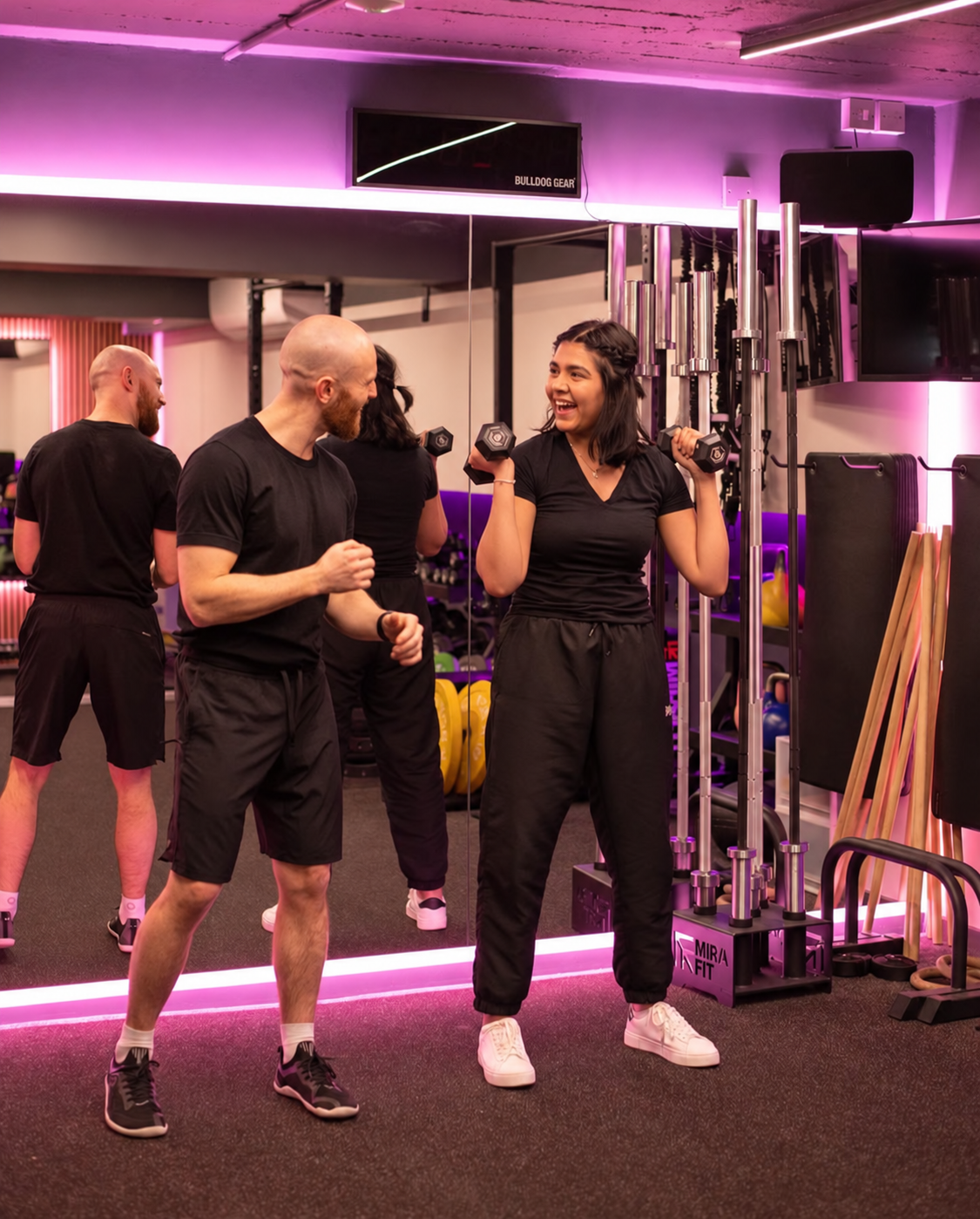 A group of people at a gym, with a young woman lifting dumbbells and smiling at a man. Other individuals are visible in the background, with gym equipment around them, and pink LED lighting on the ceiling.