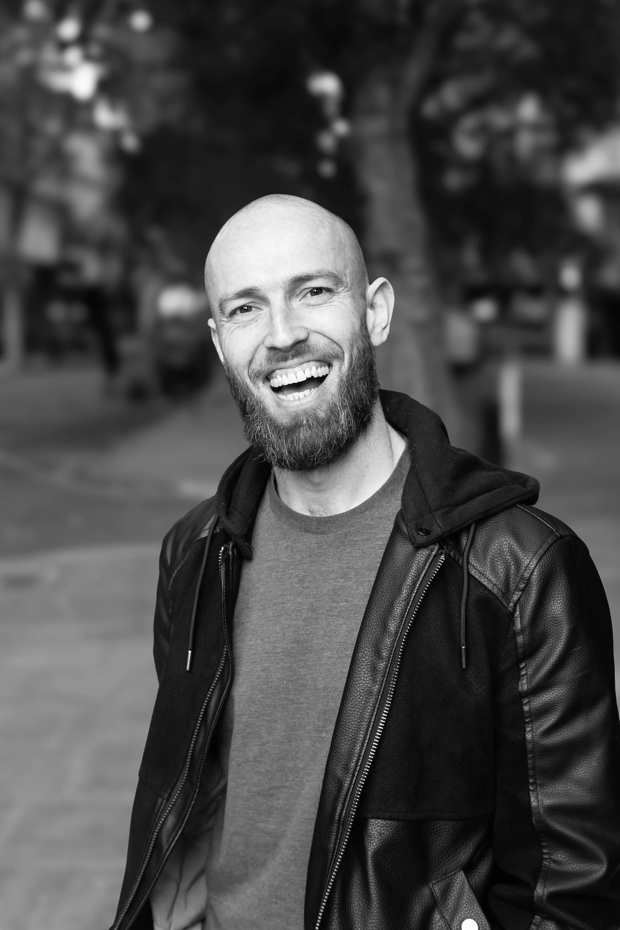 A smiling man with a beard and a shaved head, wearing a leather jacket and a T-shirt, standing outdoors in front of blurred trees in a black and white photo.
