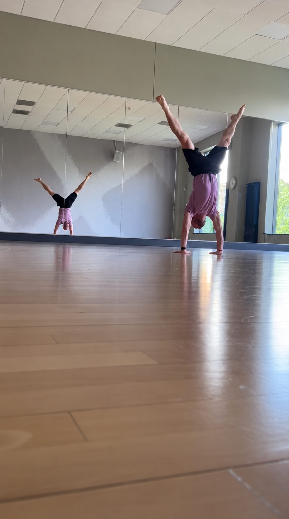 Person performing a handstand in a dance studio, visible in a large mirror, with a wooden floor and natural light from windows.