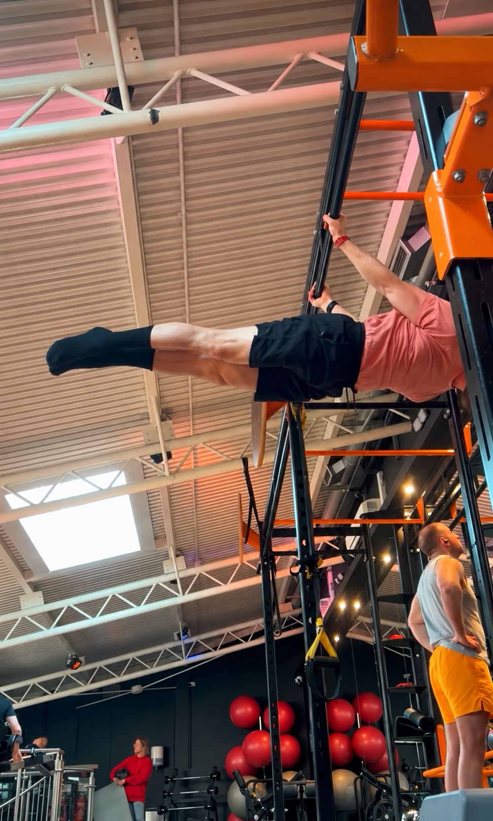 A man hanging upside down from a gym apparatus in a gym, wearing black shorts and a pink shirt, with gym equipment and other people in the background.