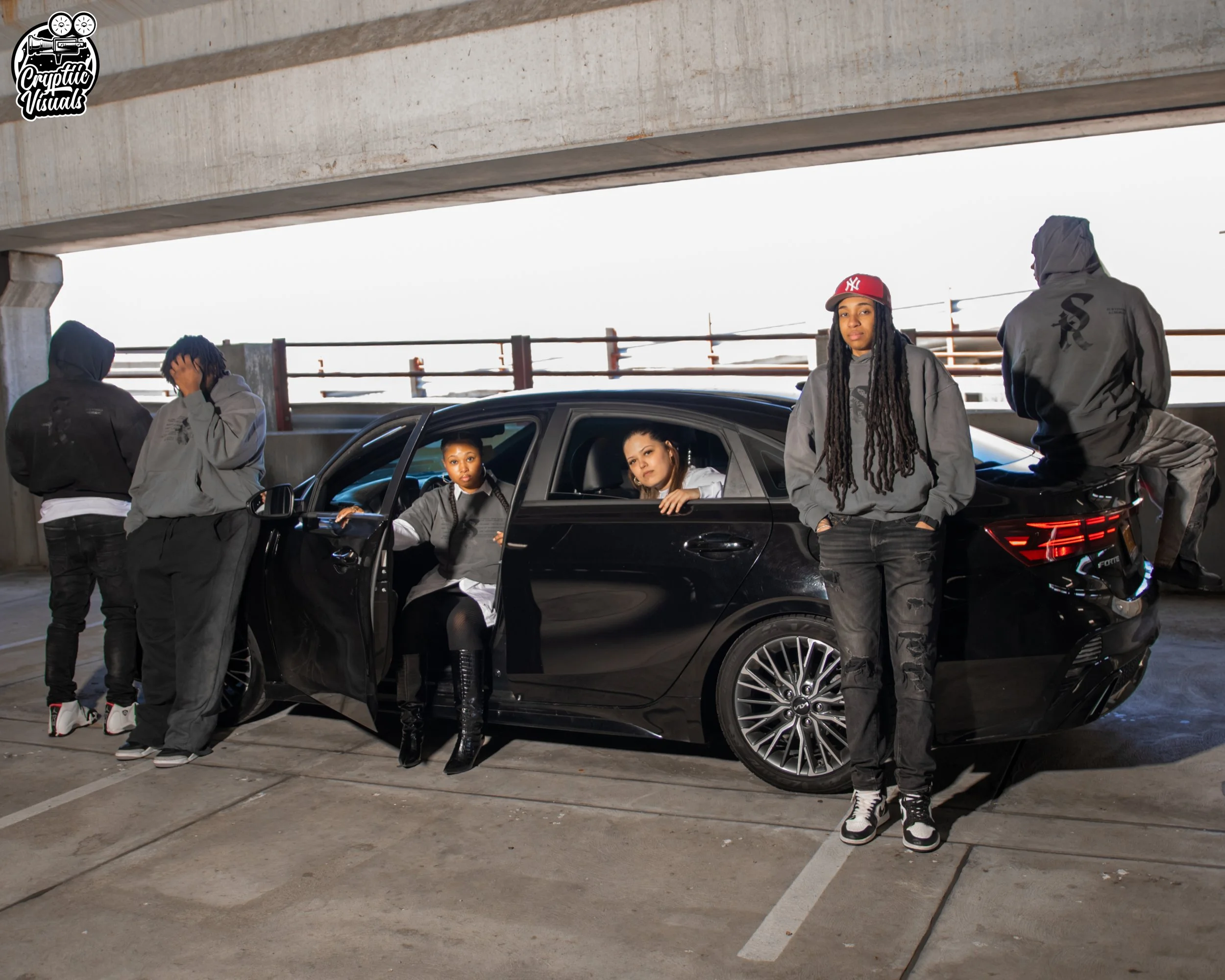 Group of young individuals in streetwear clothing standing and sitting around a black car in an urban parking garage.