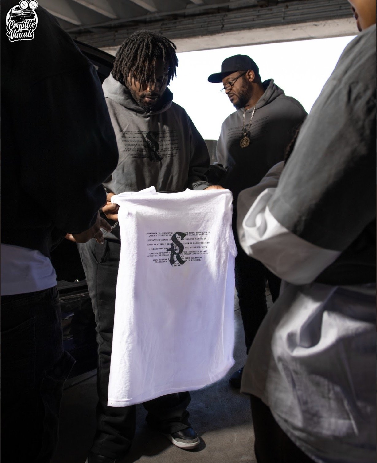 Several people are gathered around a white T-shirt with a printed design, looking at it closely. The scene appears to be outdoors under a bridge or overpass.