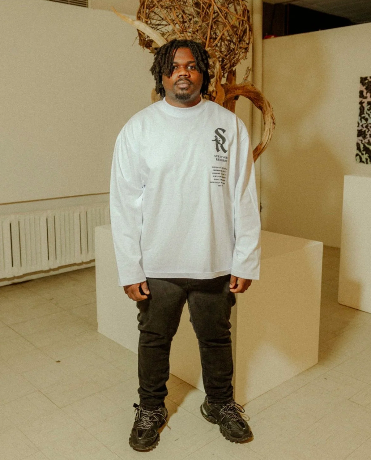 A man with dreadlocks wearing a white long-sleeve shirt, black pants, and hiking shoes stands indoors in front of abstract art and sculptures.