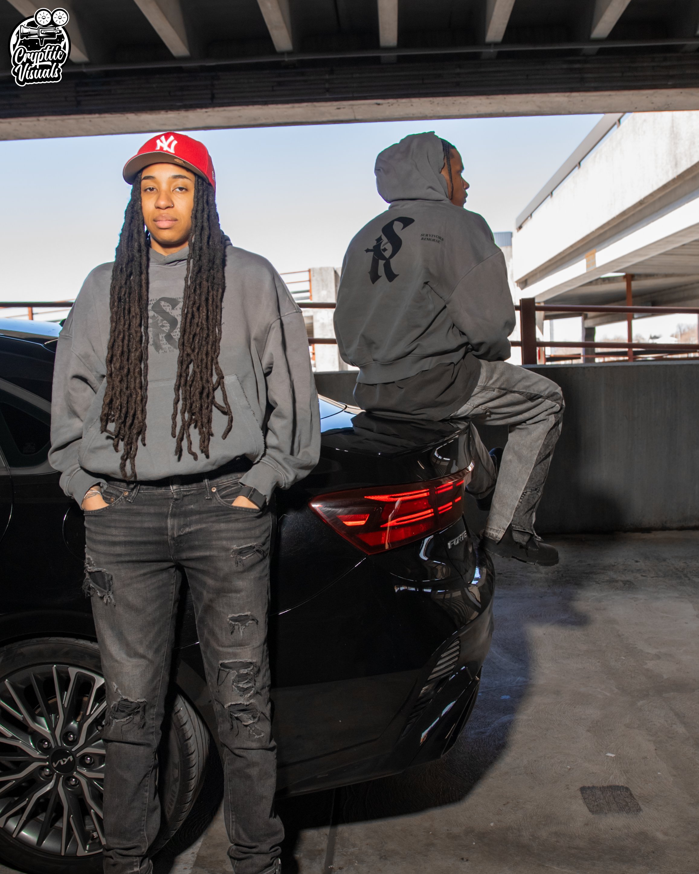 Two young men in casual clothing pose under a parking garage structure with a black car behind them. One is standing and facing forward, wearing a red New York Yankees cap and a gray hoodie. The other is sitting on the car, facing away, wearing a gray hoodie with black lettering and black jeans.
