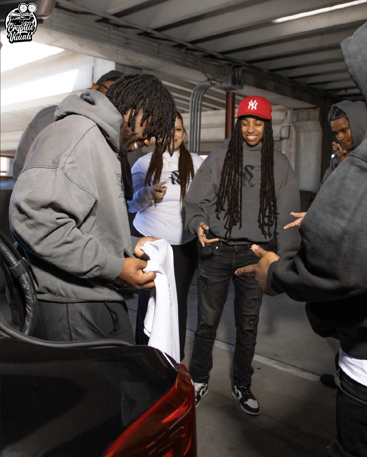 Group of young adults in a parking garage, gathered around and engaged in conversation, smiling and laughing.