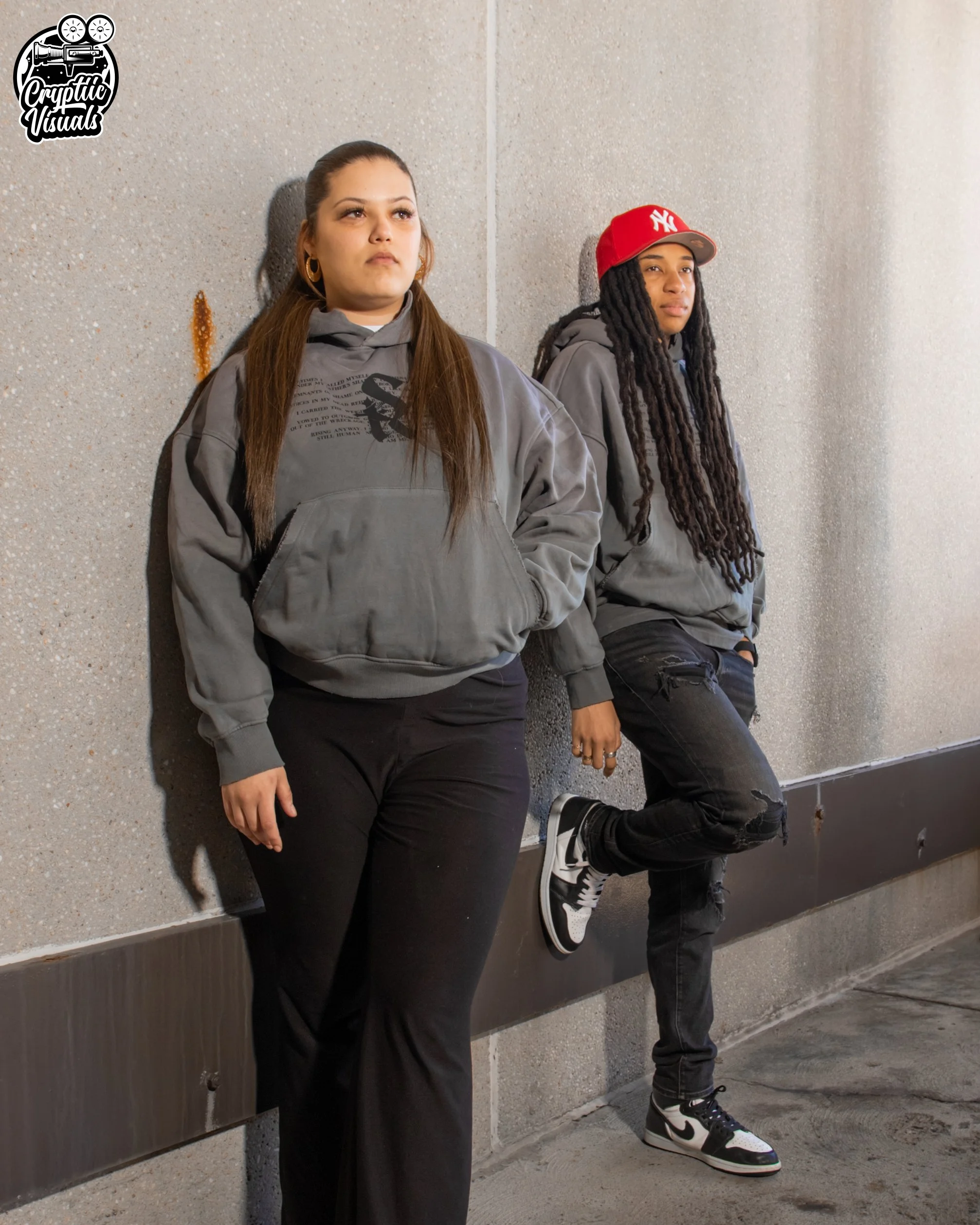 Two young women leaning against a speckled gray wall with confident expressions. The woman on the left has long brown hair, gold hoop earrings, and is wearing a gray hoodie and black pants. The woman on the right has long dreadlocks, a red baseball cap with a white logo, and a gray hoodie. She is wearing black jeans with ripped knees and black-and-white sneakers.
