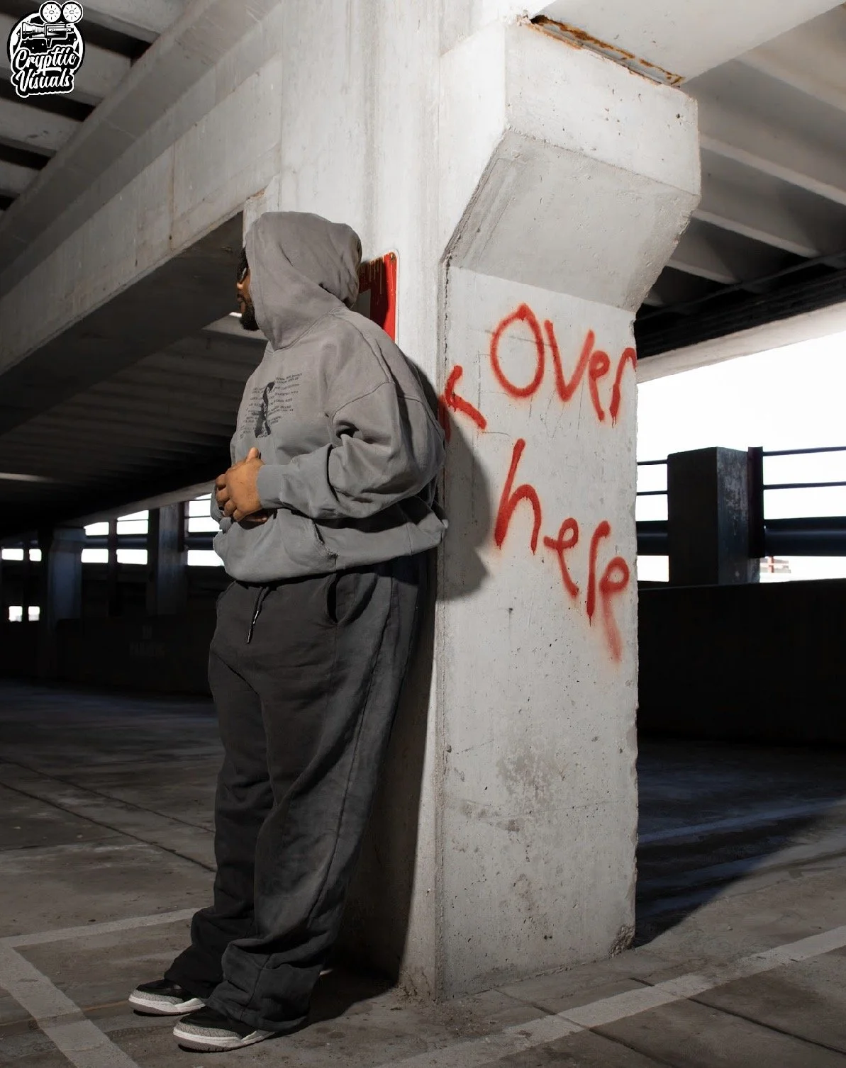 A man in a gray hoodie and gray sweatpants leaning against a concrete pillar with red graffiti reading "OVER HERE" in an indoor parking garage.