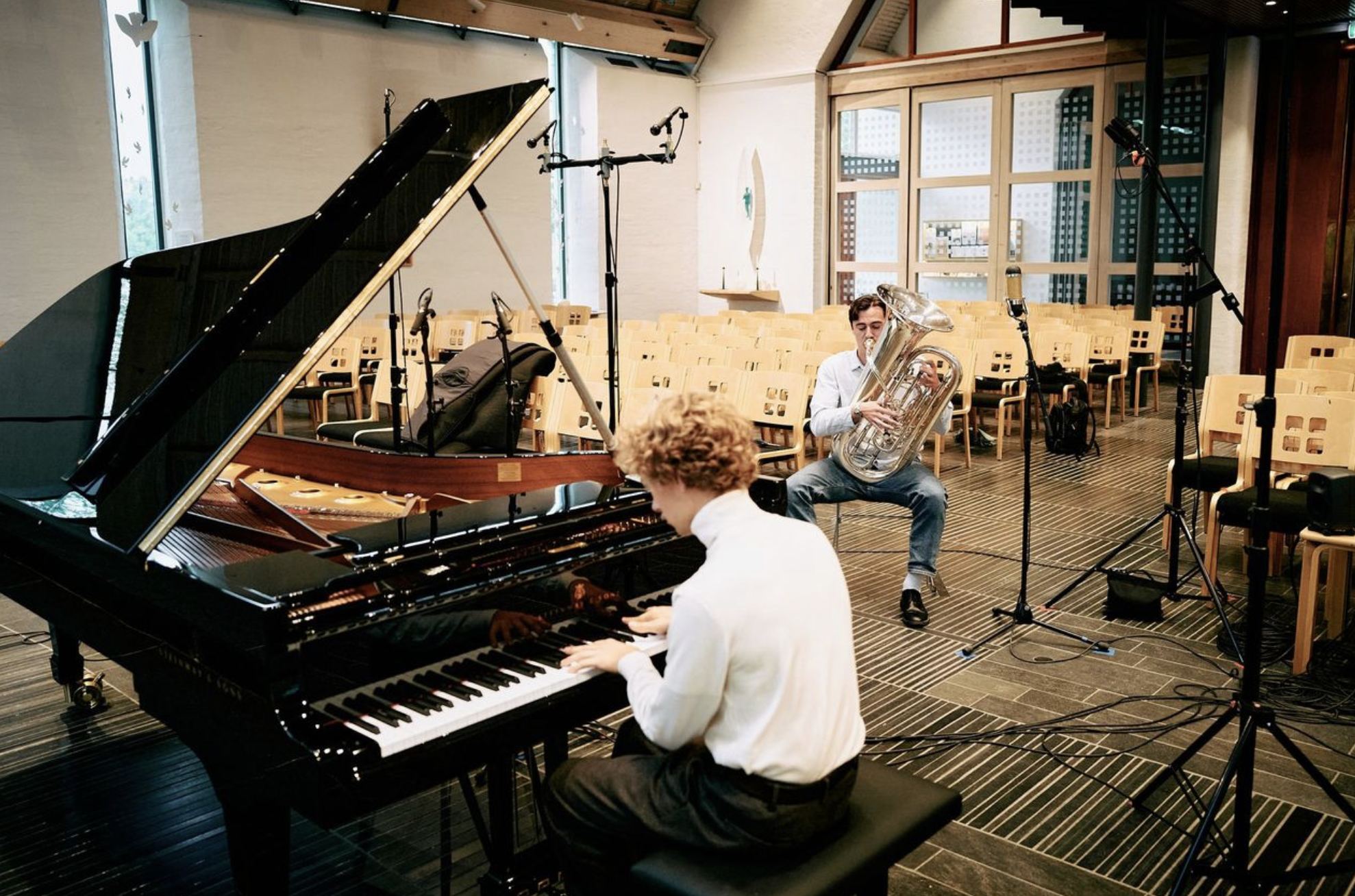 A person playing a grand piano, another person playing a euphonium, and a person playing a trombone inside a music hall with empty chairs and microphones.