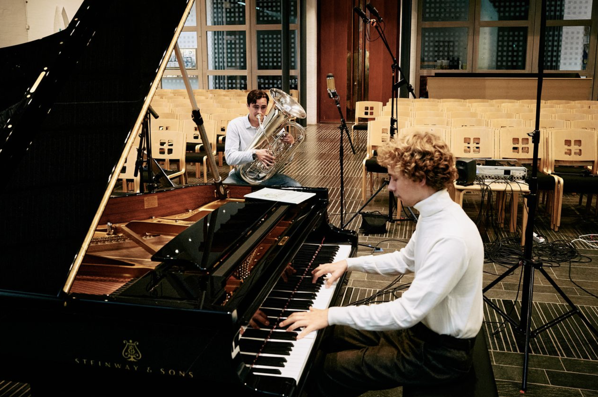 Two musicians performing in a room with wooden chairs and a grand piano. One musician is playing the piano, and the other is playing a tuba. The room has large windows, and recording equipment is set up around the performers.