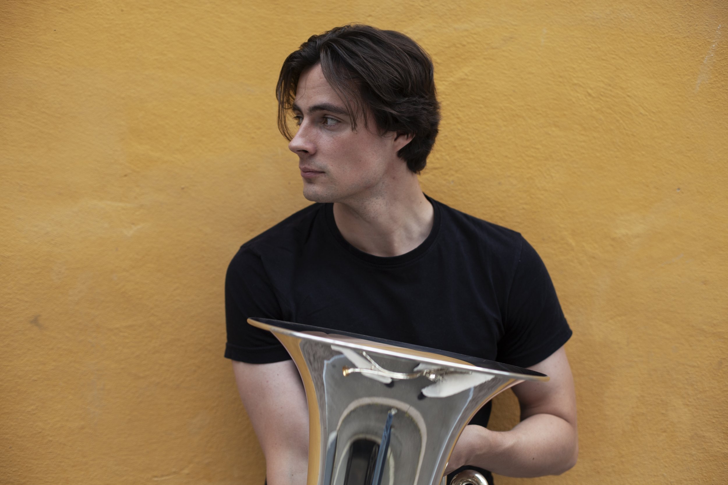 A young man with brown hair wearing a black shirt holding and looking at a skateboard with a yellow wall in the background.