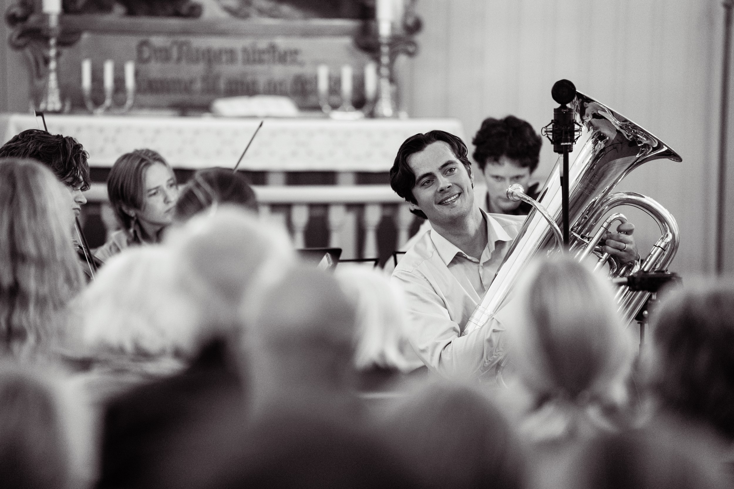 A black and white photo of a young man playing a tuba during a musical performance, surrounded by an audience.
