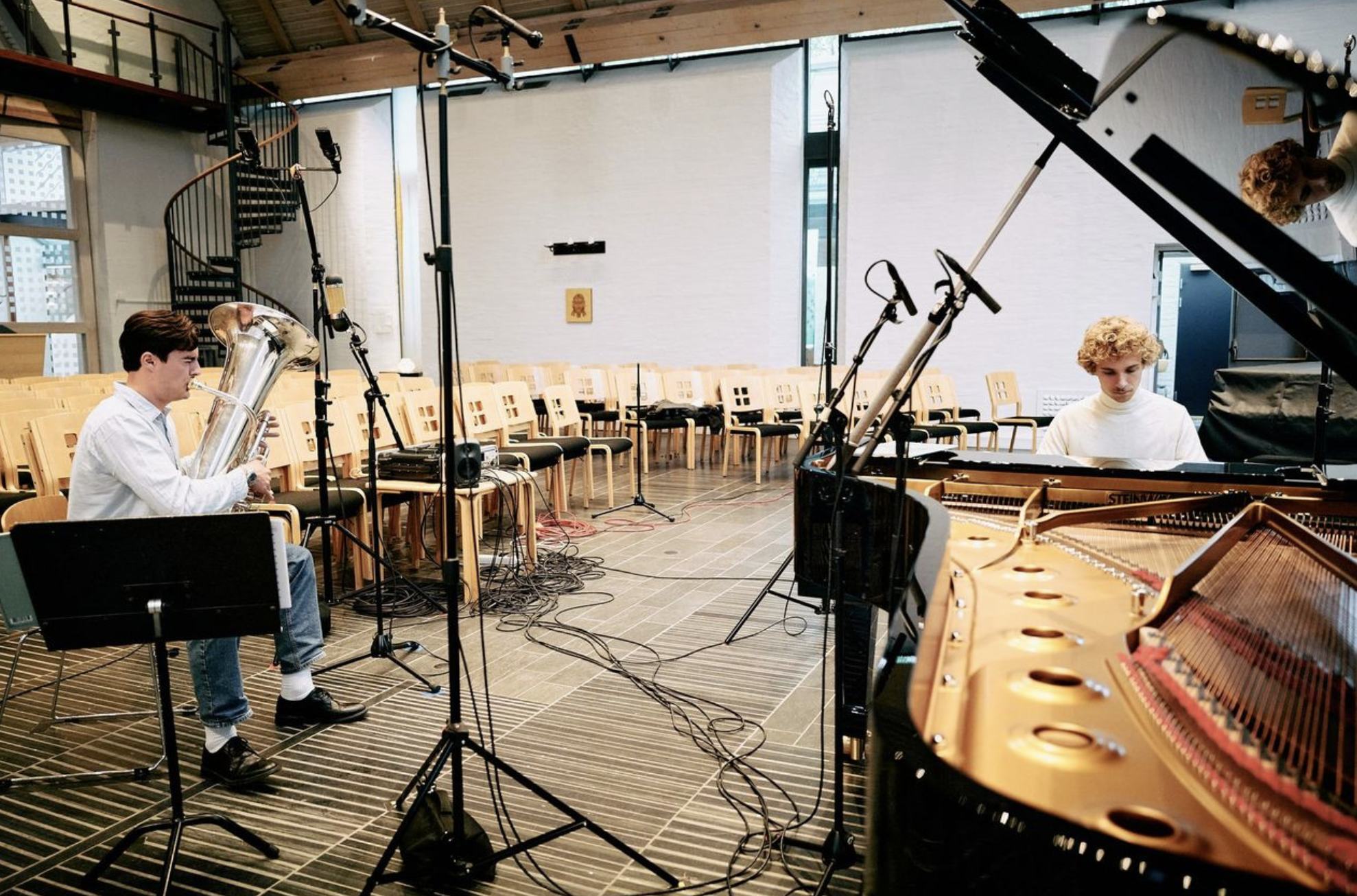 Two musicians playing instruments in a music rehearsal space with chairs, microphones, and a grand piano.