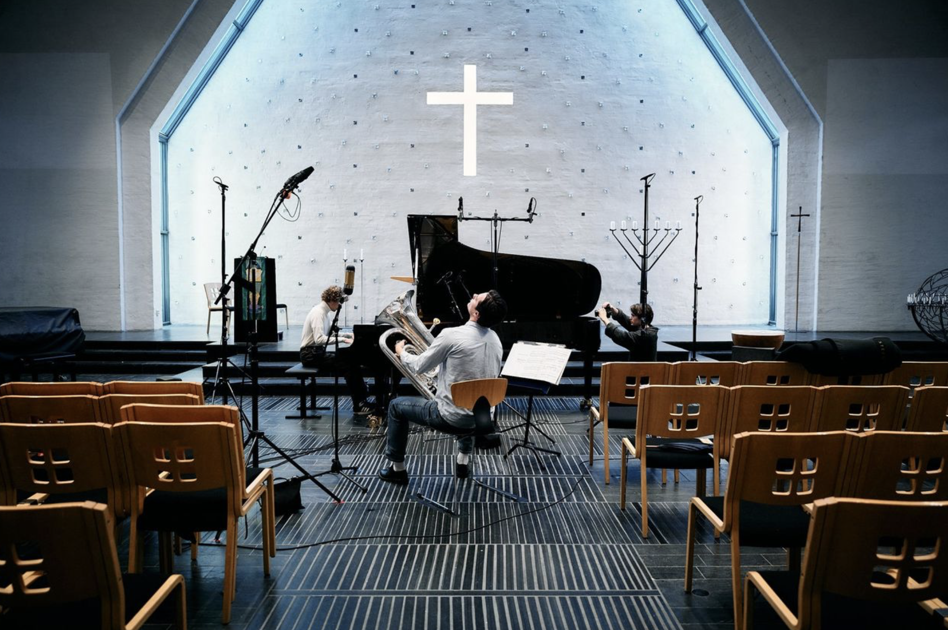 Inside a church or concert hall with musicians preparing for a performance. There is a grand piano, music stands, and musicians tuning or arranging instruments. The background features a large cross illuminated on a white wall, indicating a religious