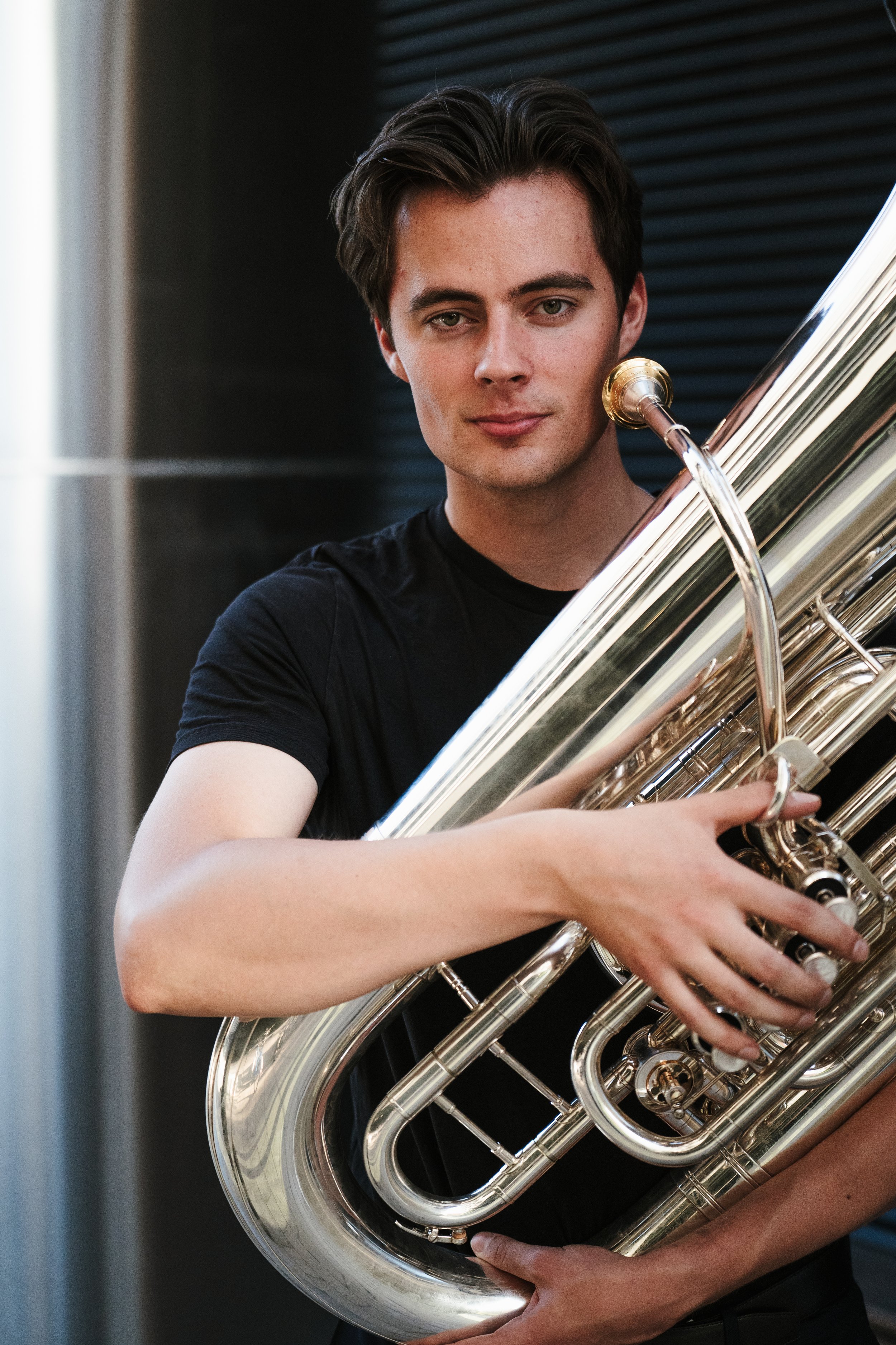 Young man holding a shiny silver euphonium in a room with dark background.