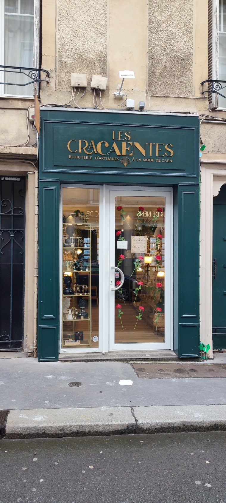 Façade d'une boutique nommée 'Les Cracaentes', spécialisée en bijouterie artisanale à la mode de Caen. La vitrine est décorée de roses et la porte est en verre.