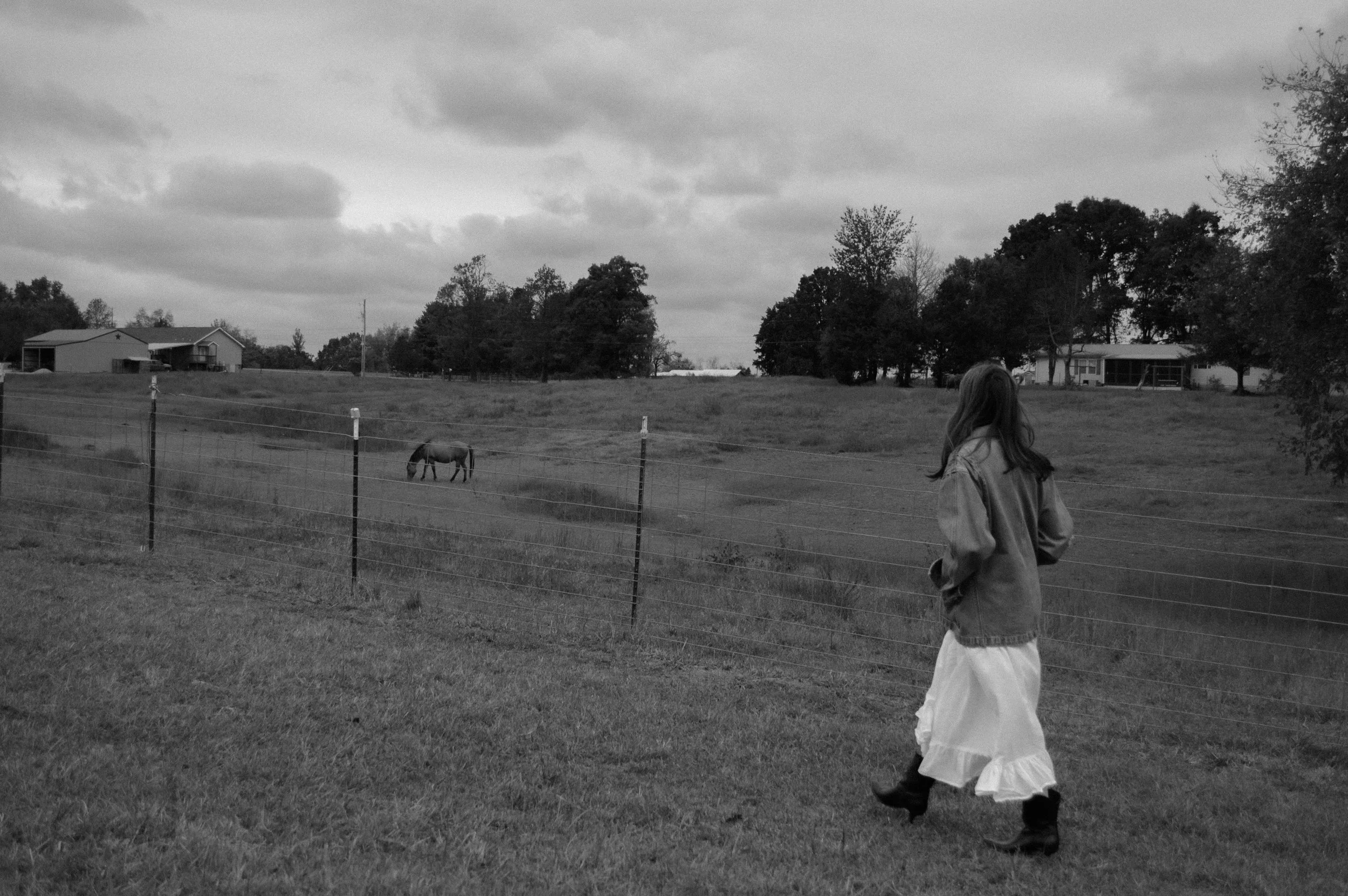 A girl in a long dress and jacket walking on a grassy field past a wooden fence, with houses and trees in the background, and a cow grazing near the fence on a cloudy day.