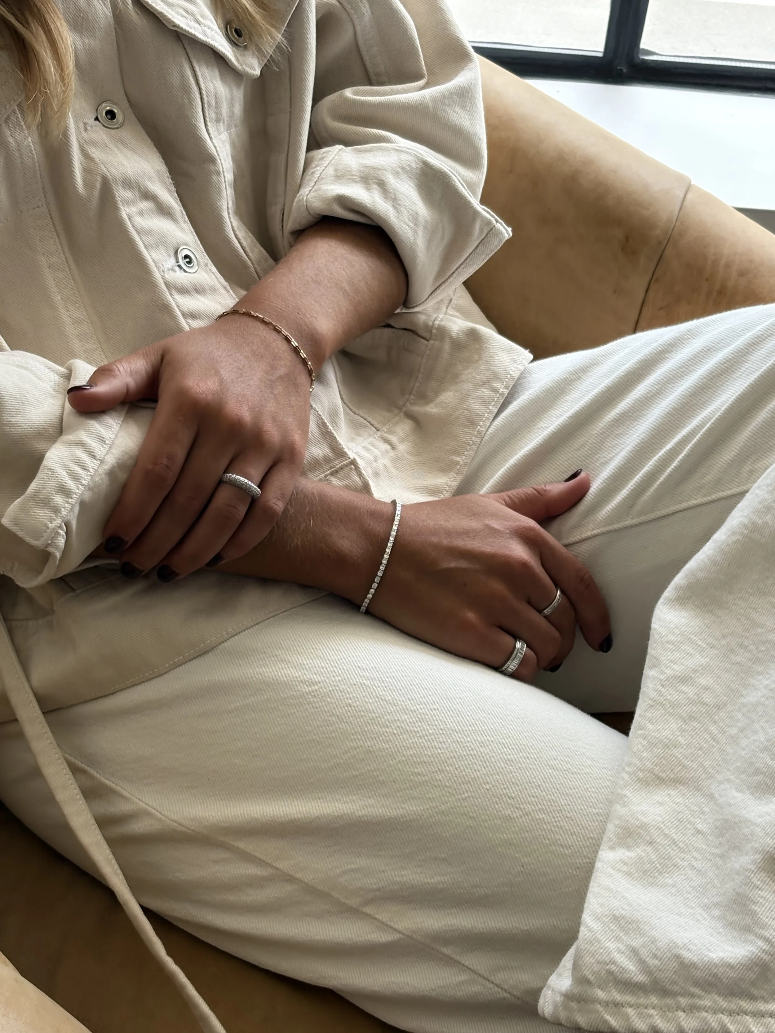 Close-up of a person sitting, wearing a beige denim jacket and beige pants, with their hands resting on their lap, showing rings and bracelets.