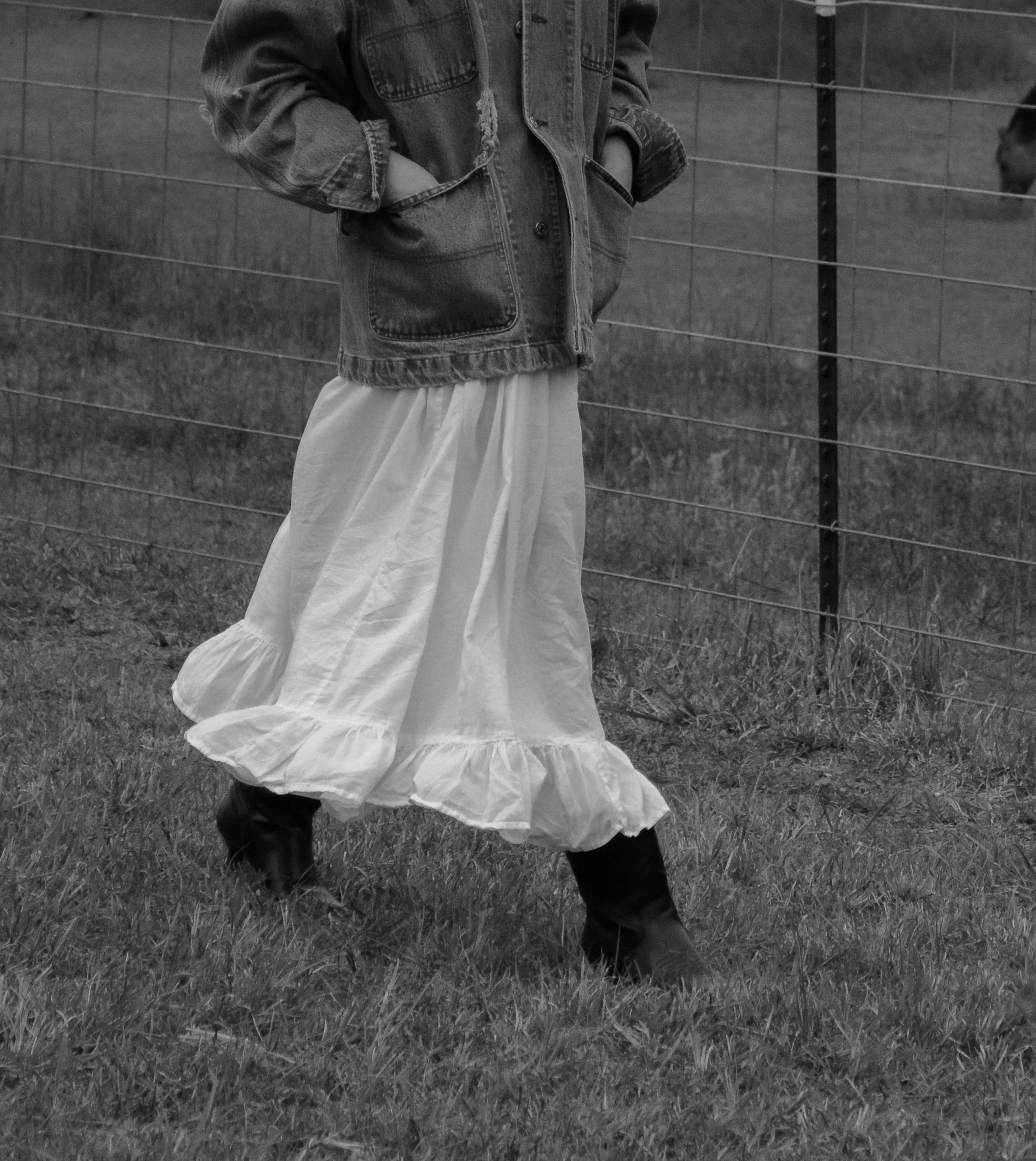 A person standing outdoors on grass, wearing a denim jacket and a long, flowing white skirt, with hands in pockets, near a wire fence.