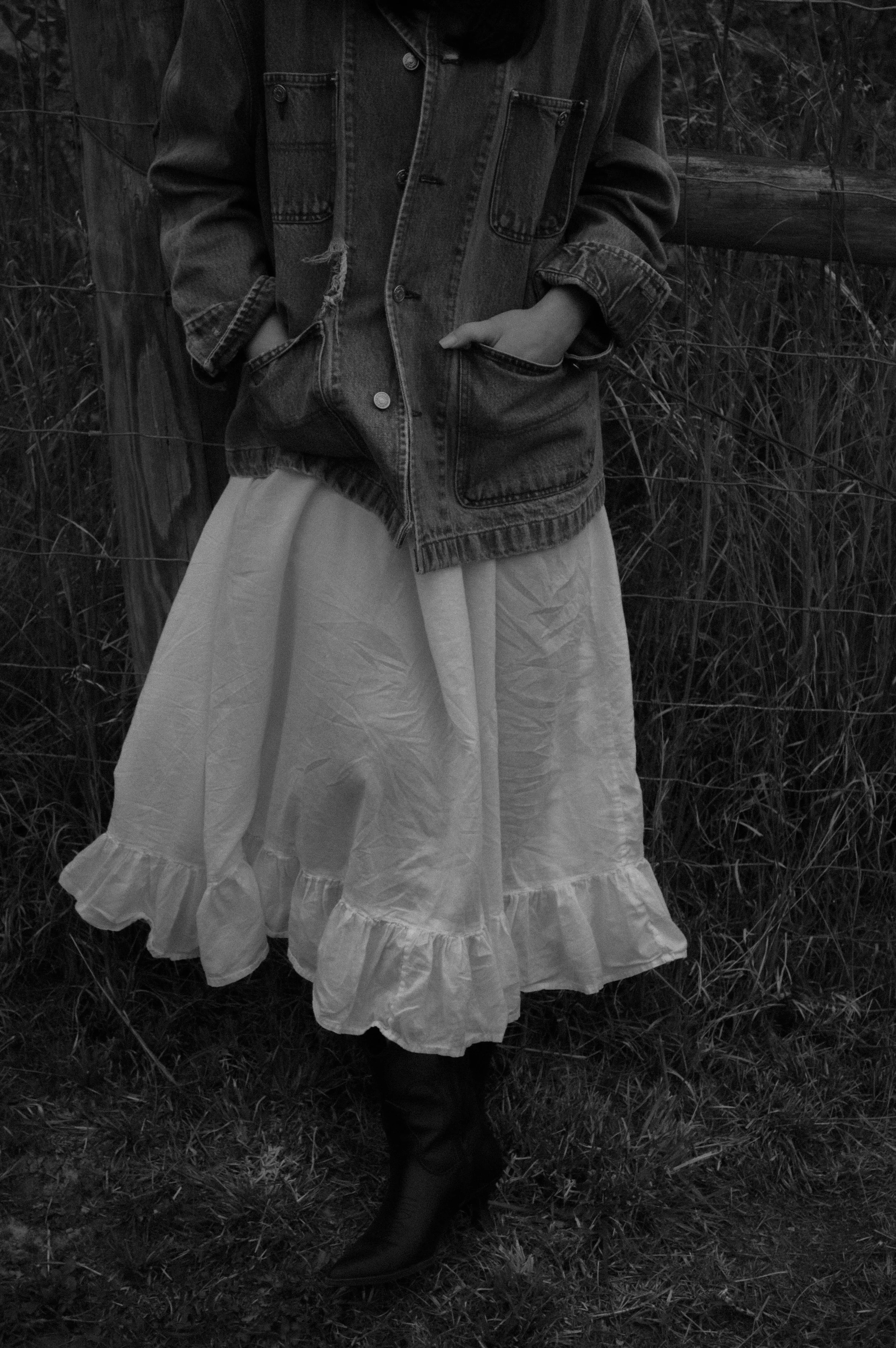 Person wearing a denim jacket and a long layered skirt standing outdoors near a wooden fence with tall grass.