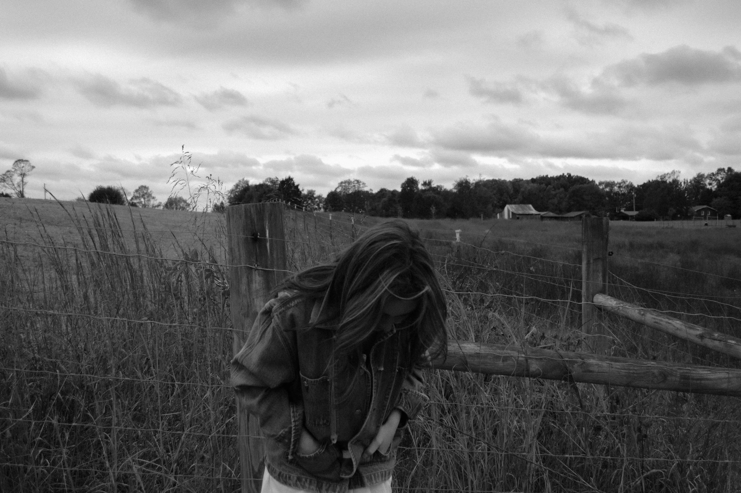 A person with long hair, wearing a jacket, stands near a rustic wooden fence in an open grassy field under cloudy skies, with trees and farm buildings in the background.