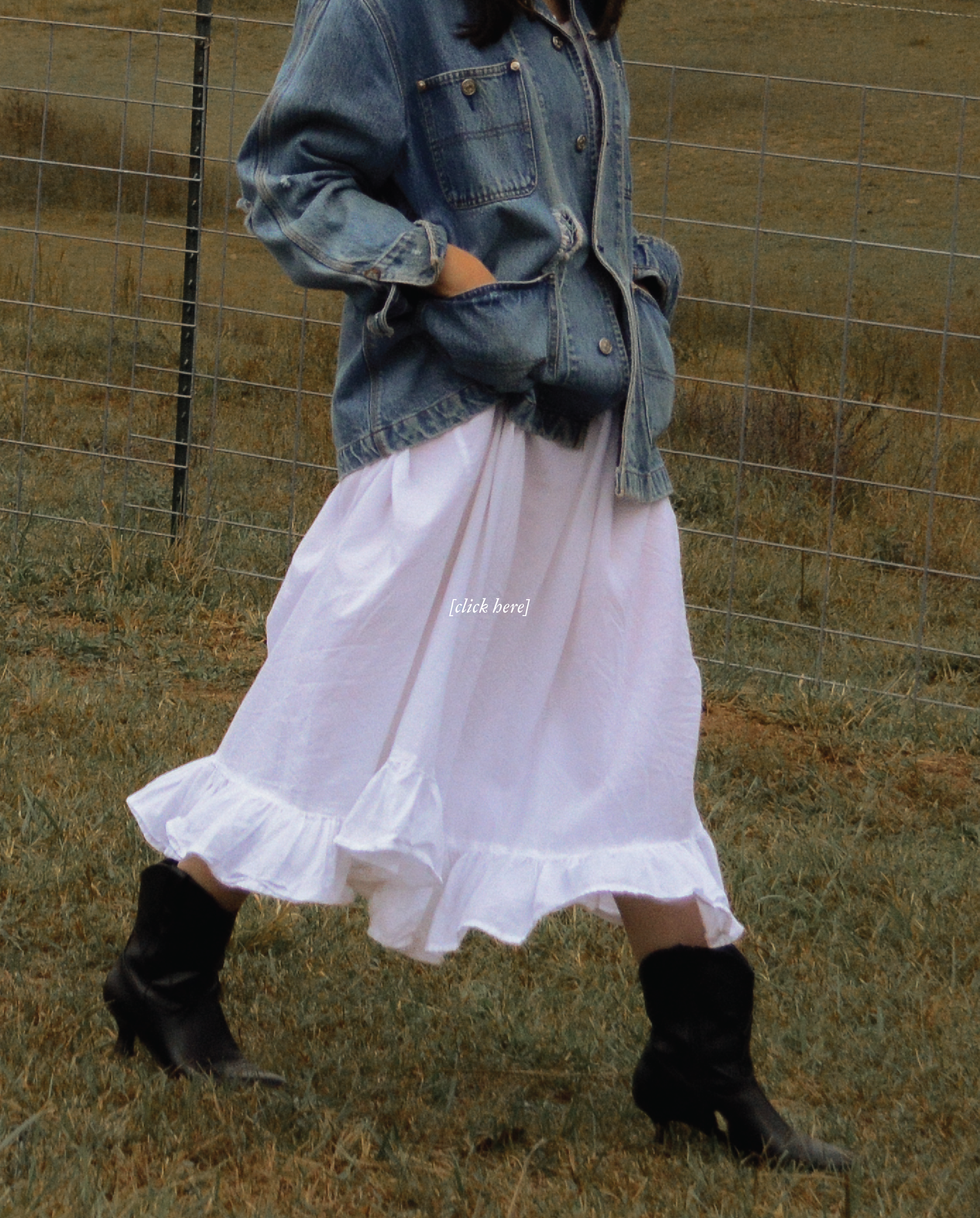A person standing outdoors near a metal fence, wearing a denim jacket, a white dress, and black boots on grassy ground.